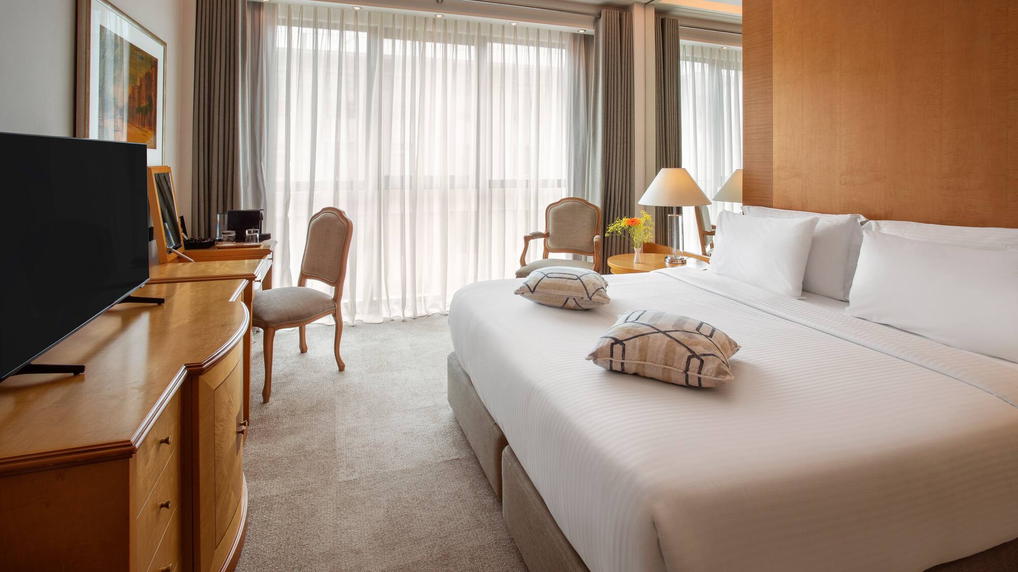 Luxury hotel room with king bed, flat-screen TV, wooden desk, and bright window.