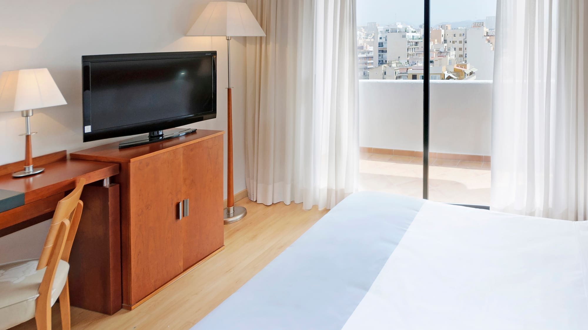 Bright hotel room with desk, TV, bed, and balcony featuring city views.