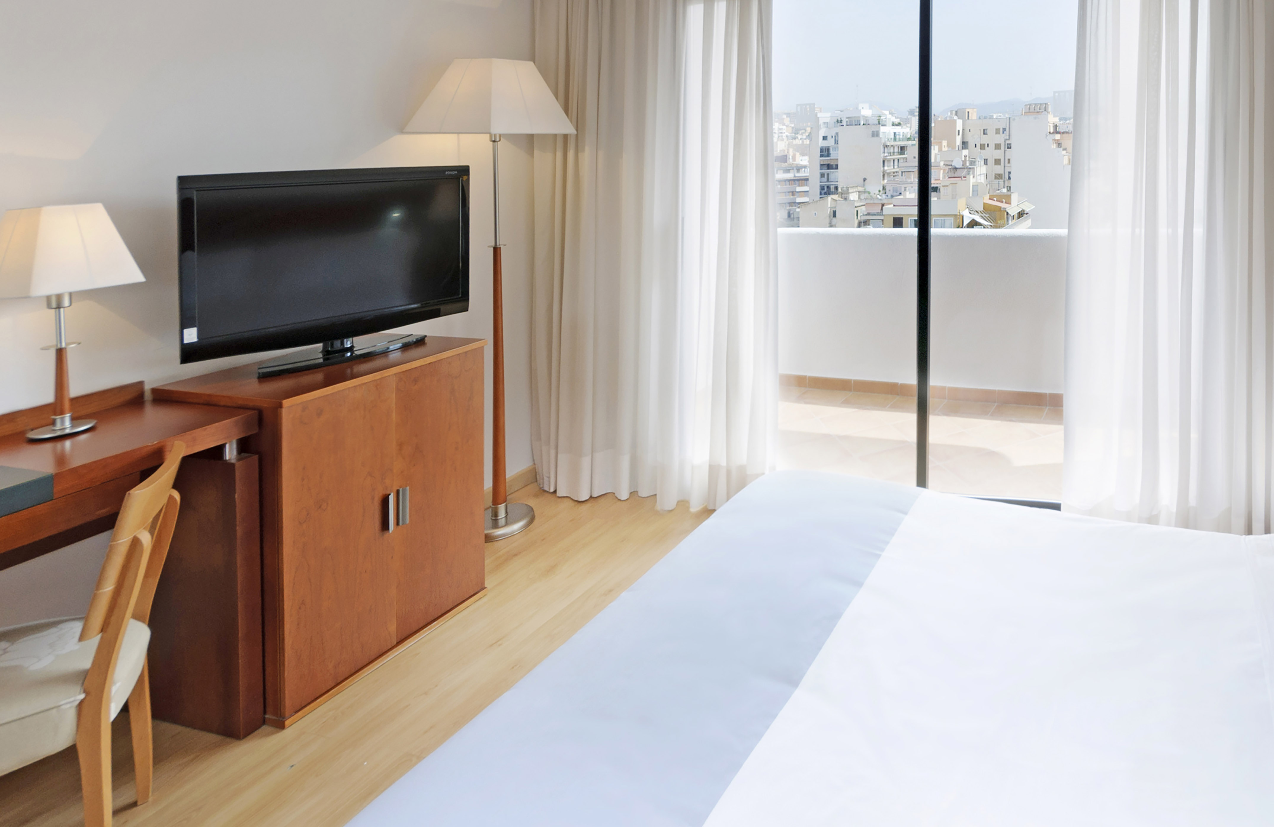 Bright hotel room with desk, TV, bed, and balcony featuring city views.