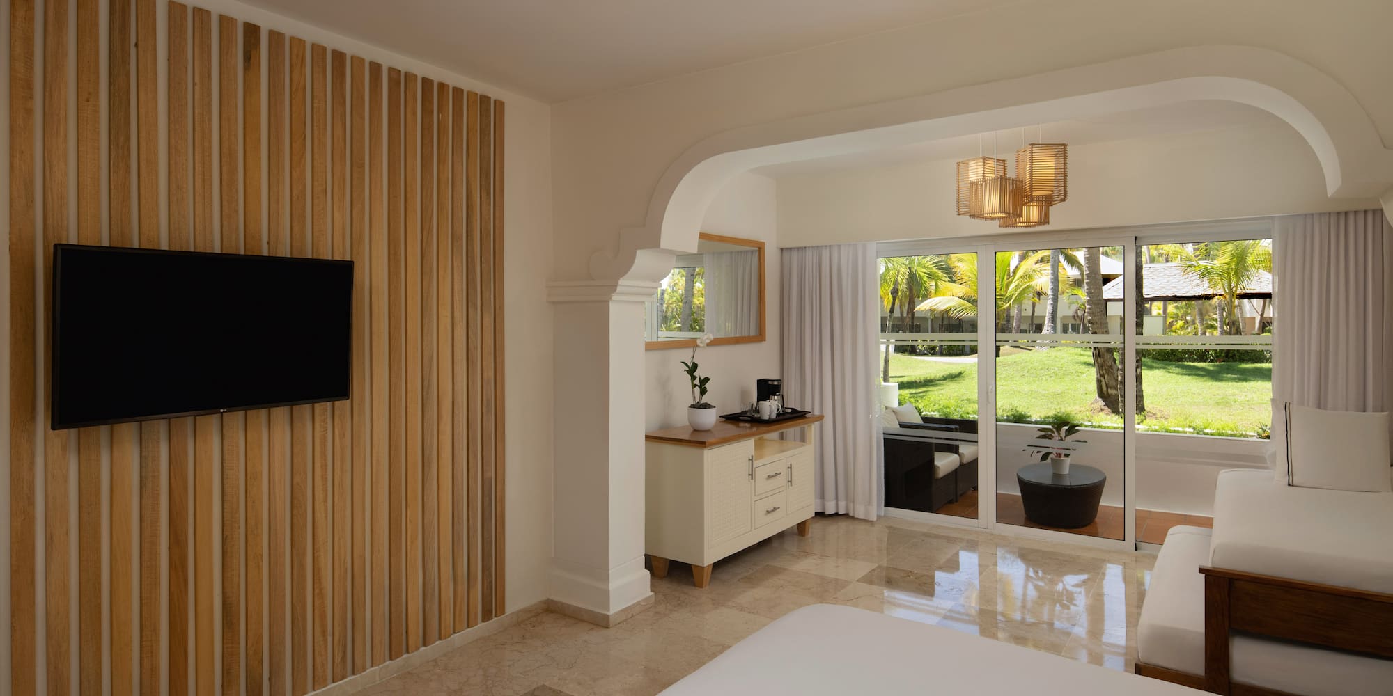 Modern resort bedroom with TV, wood accent wall, and tropical patio view.