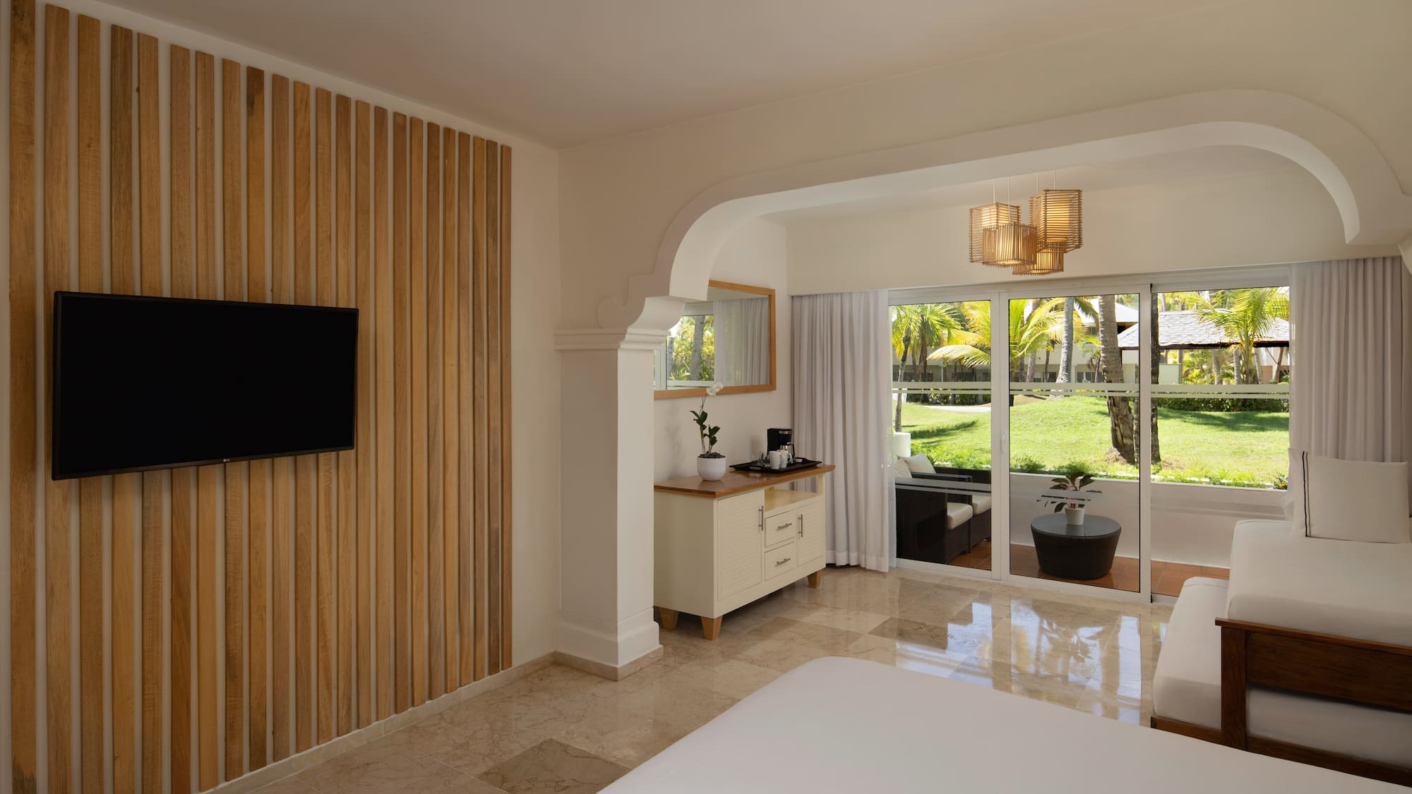 Modern resort bedroom with TV, wood accent wall, and tropical patio view.