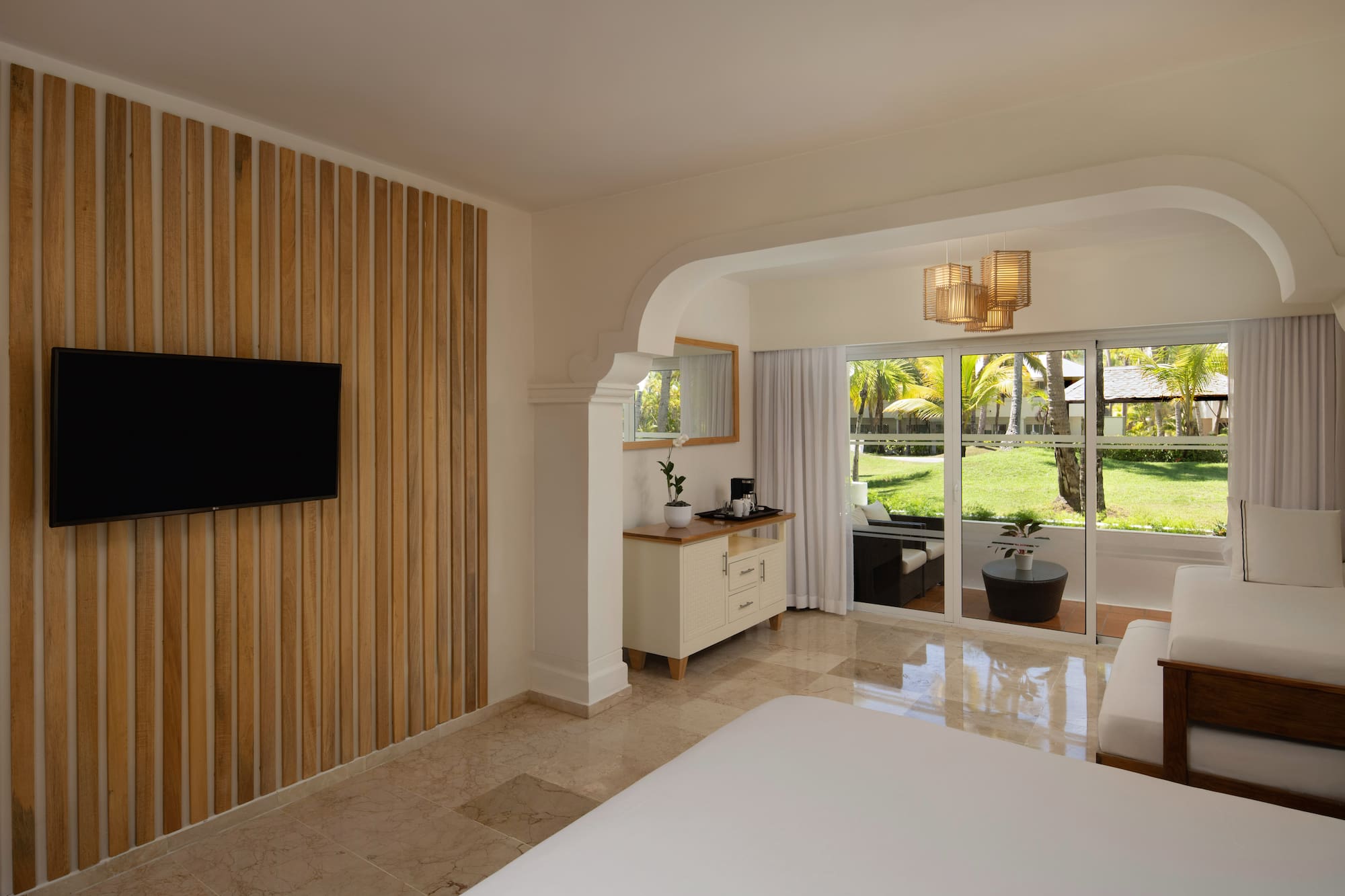 Modern resort bedroom with TV, wood accent wall, and tropical patio view.