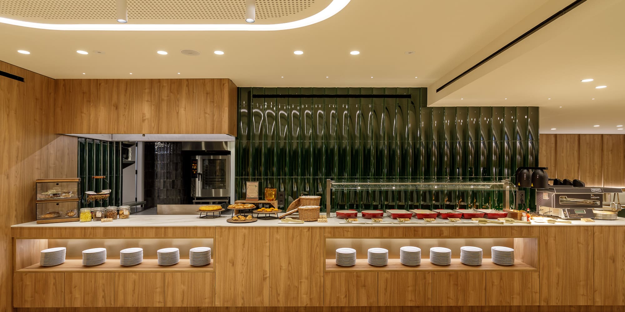 Modern hotel breakfast buffet with wooden counters, green textured wall, and prepared food.