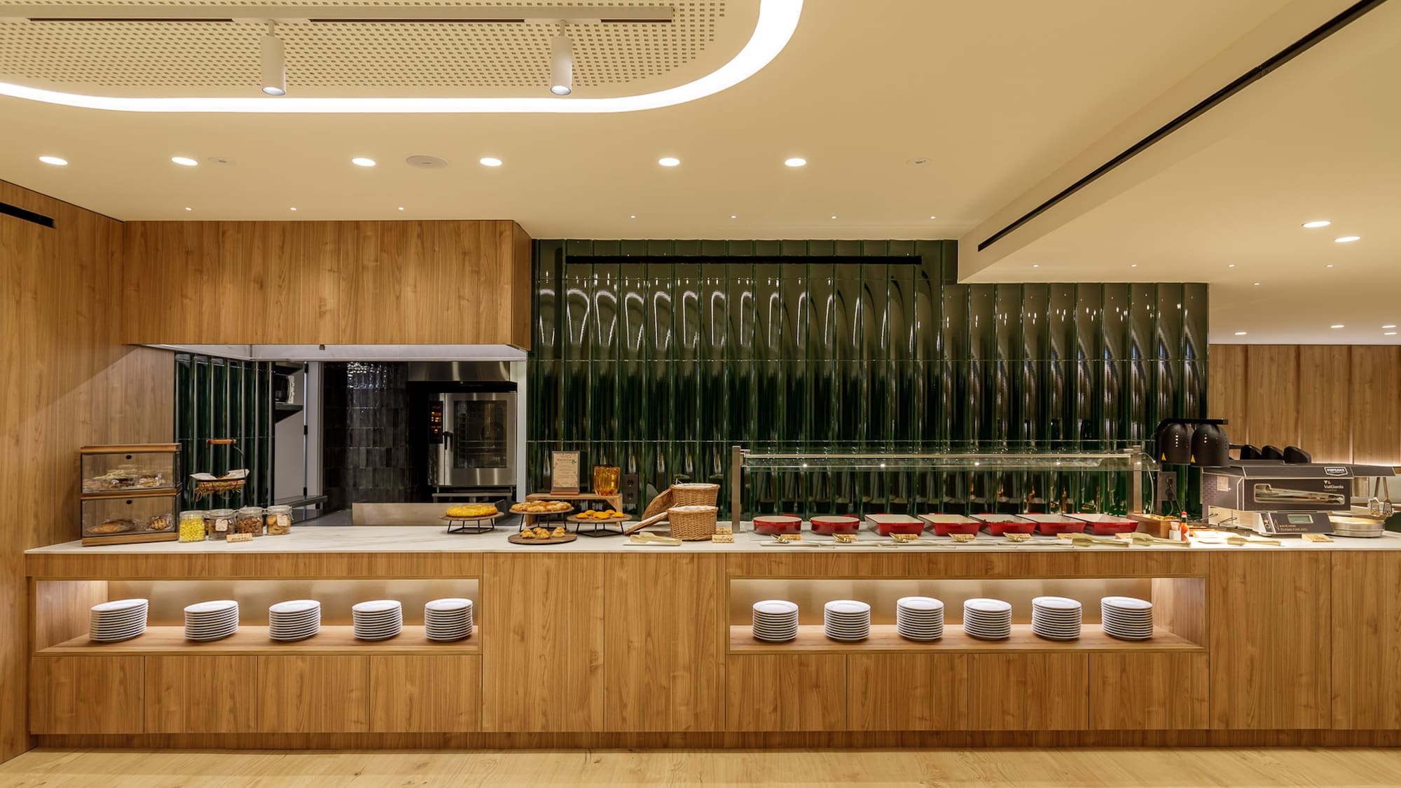 Modern hotel breakfast buffet with wooden counters, green textured wall, and prepared food.