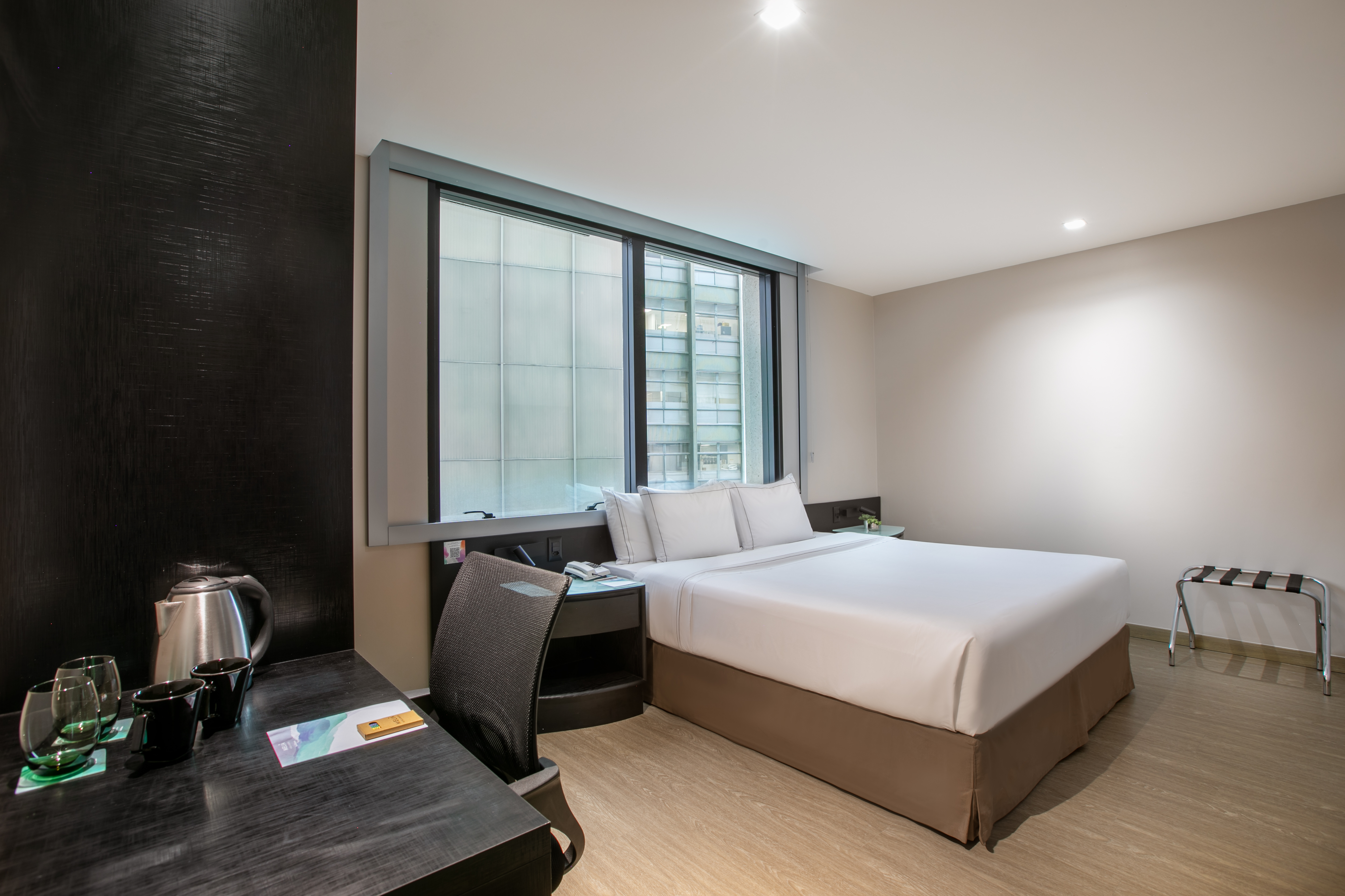 Modern hotel room with bed, desk, kettle, and large window with city view.