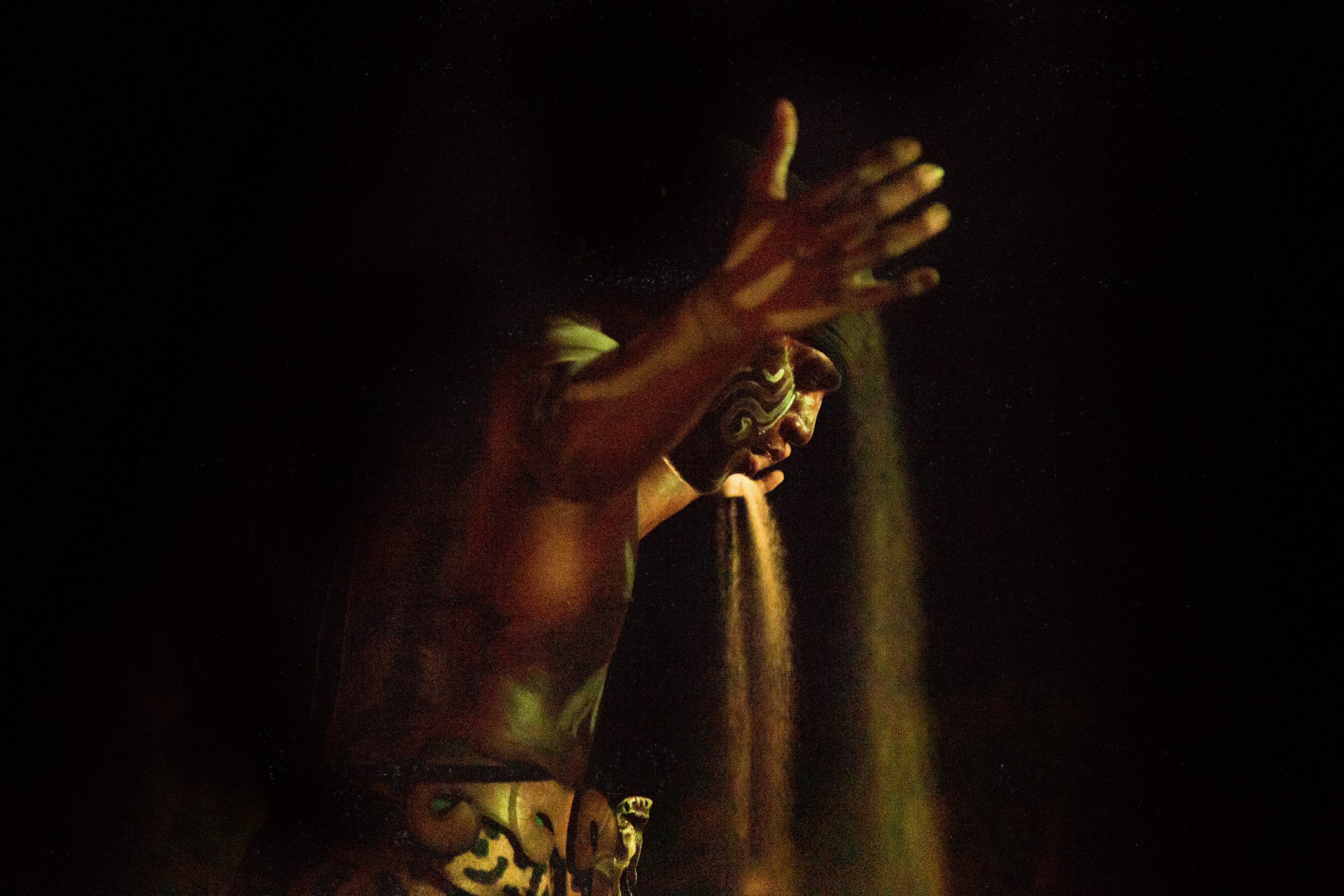 Dark figure with face paint performing sand ritual in dramatic light.