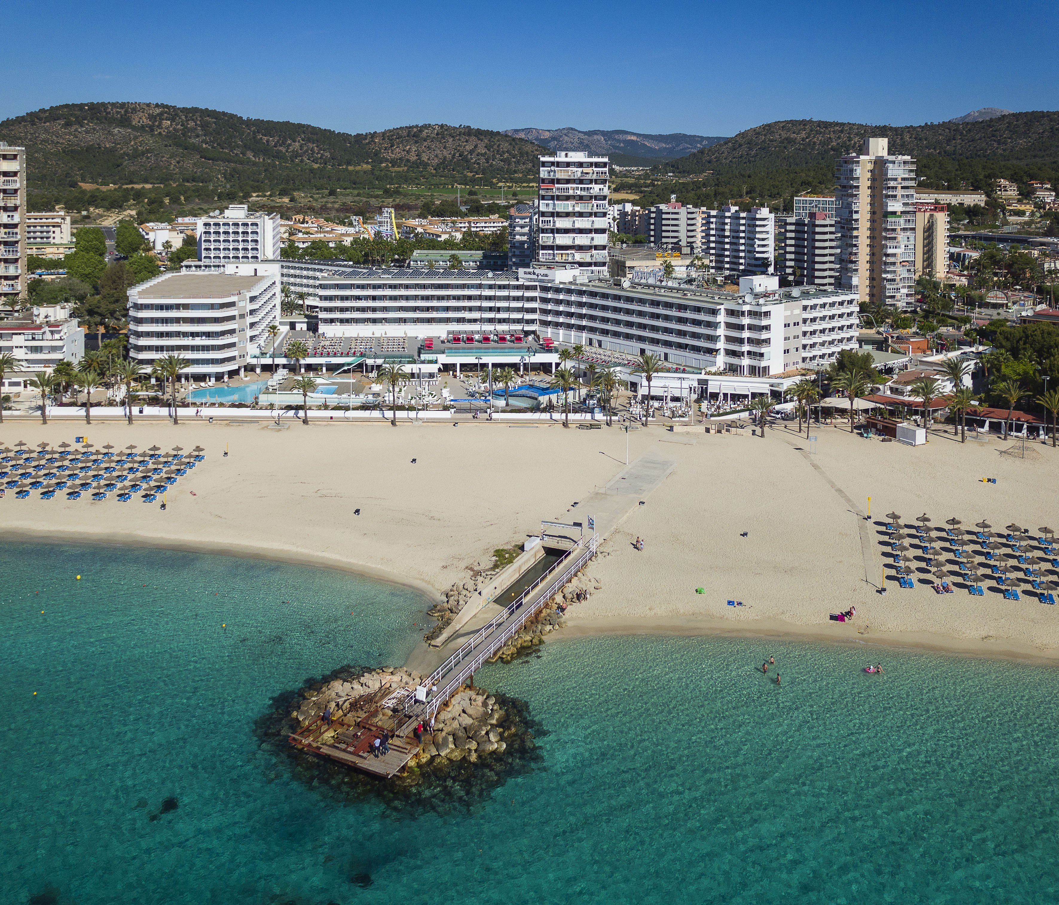 Coastal resort with hotels, sandy beach, turquoise sea, pier, mountains.