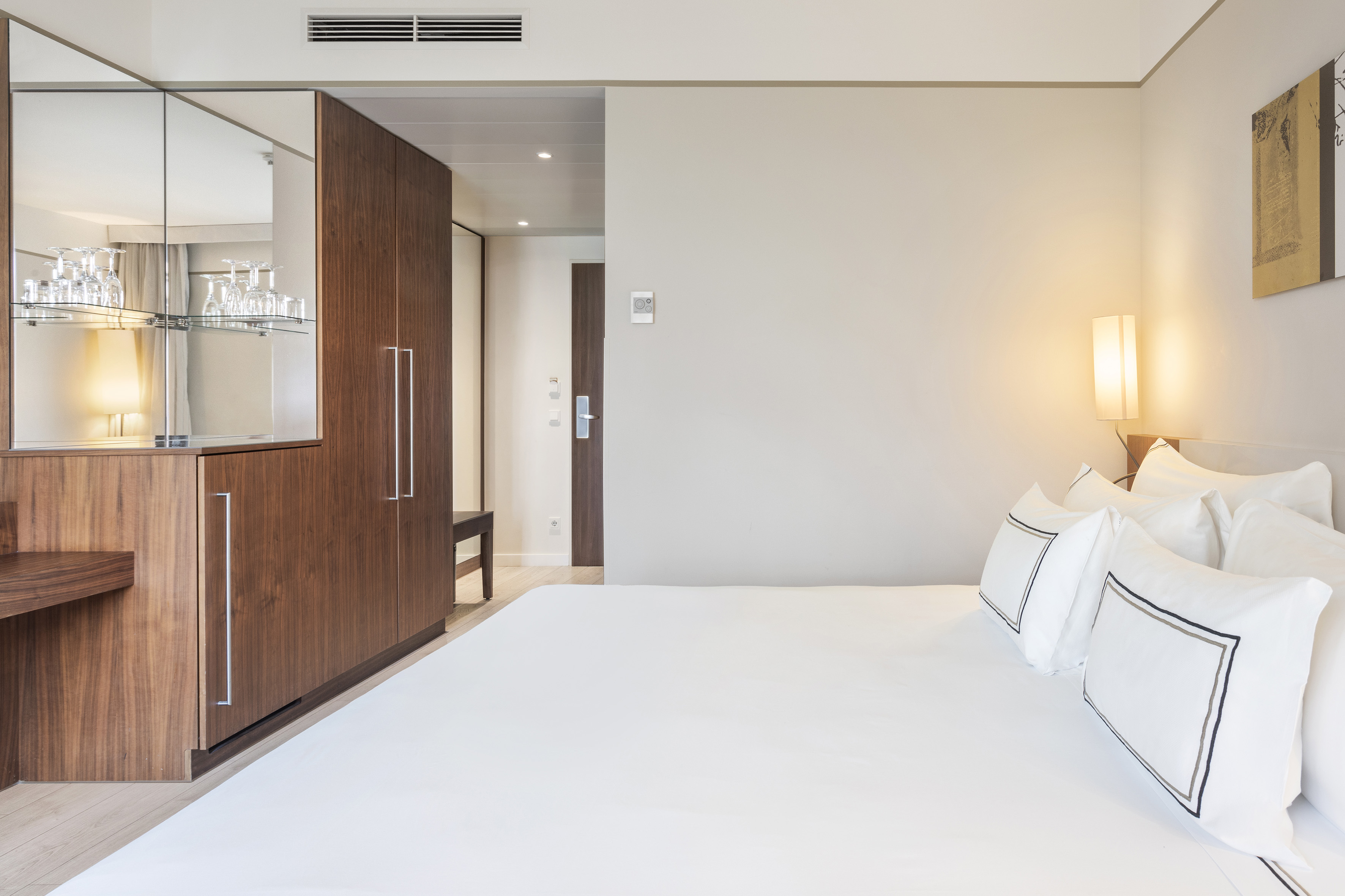Modern hotel room with large white bed, wooden wardrobe, and mirrored mini-bar.