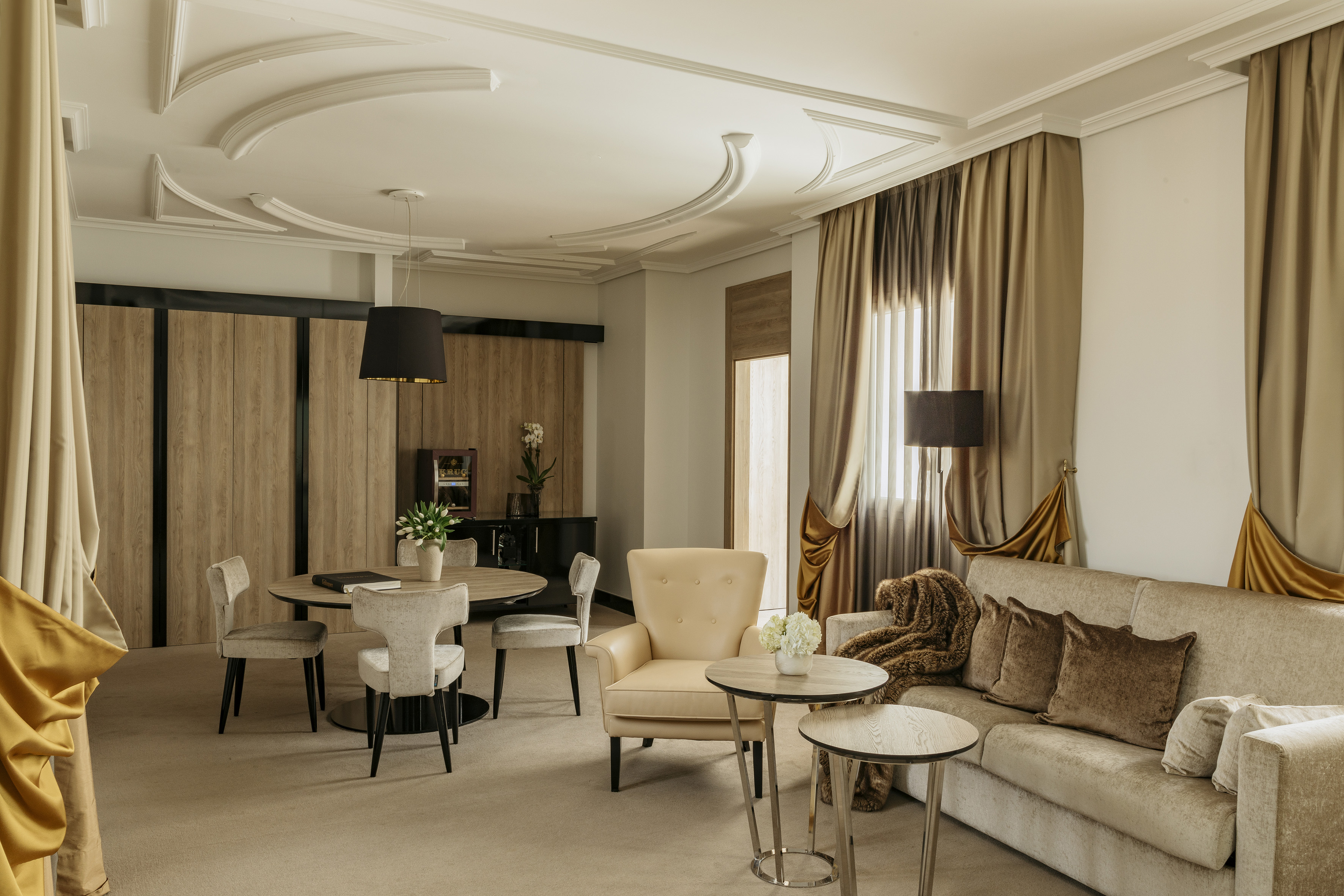 Luxury hotel suite living room with dining area, beige sofa, gold curtains.