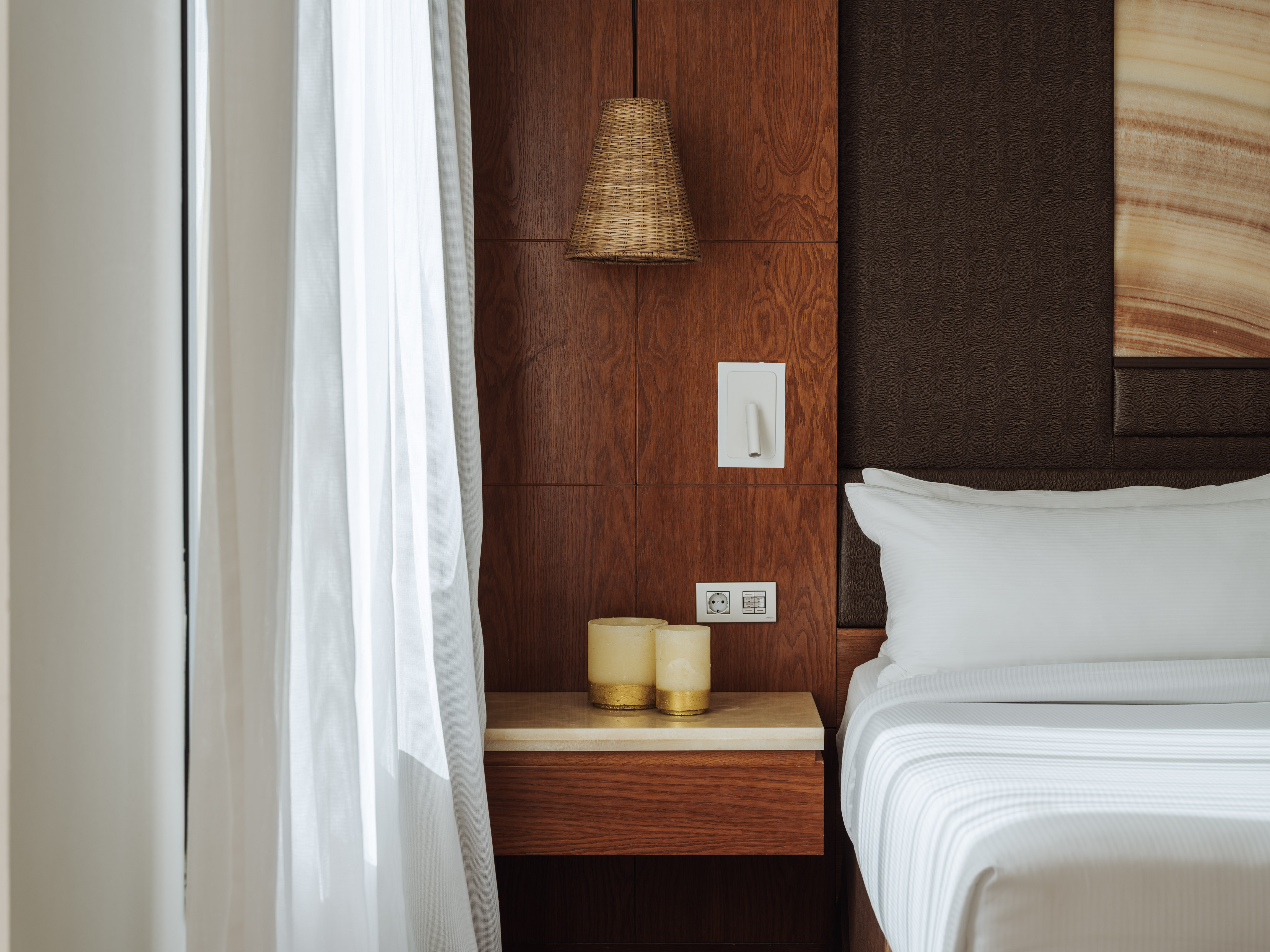 Modern hotel bedroom with wood panel, white bed, nightstand, woven lamp.
