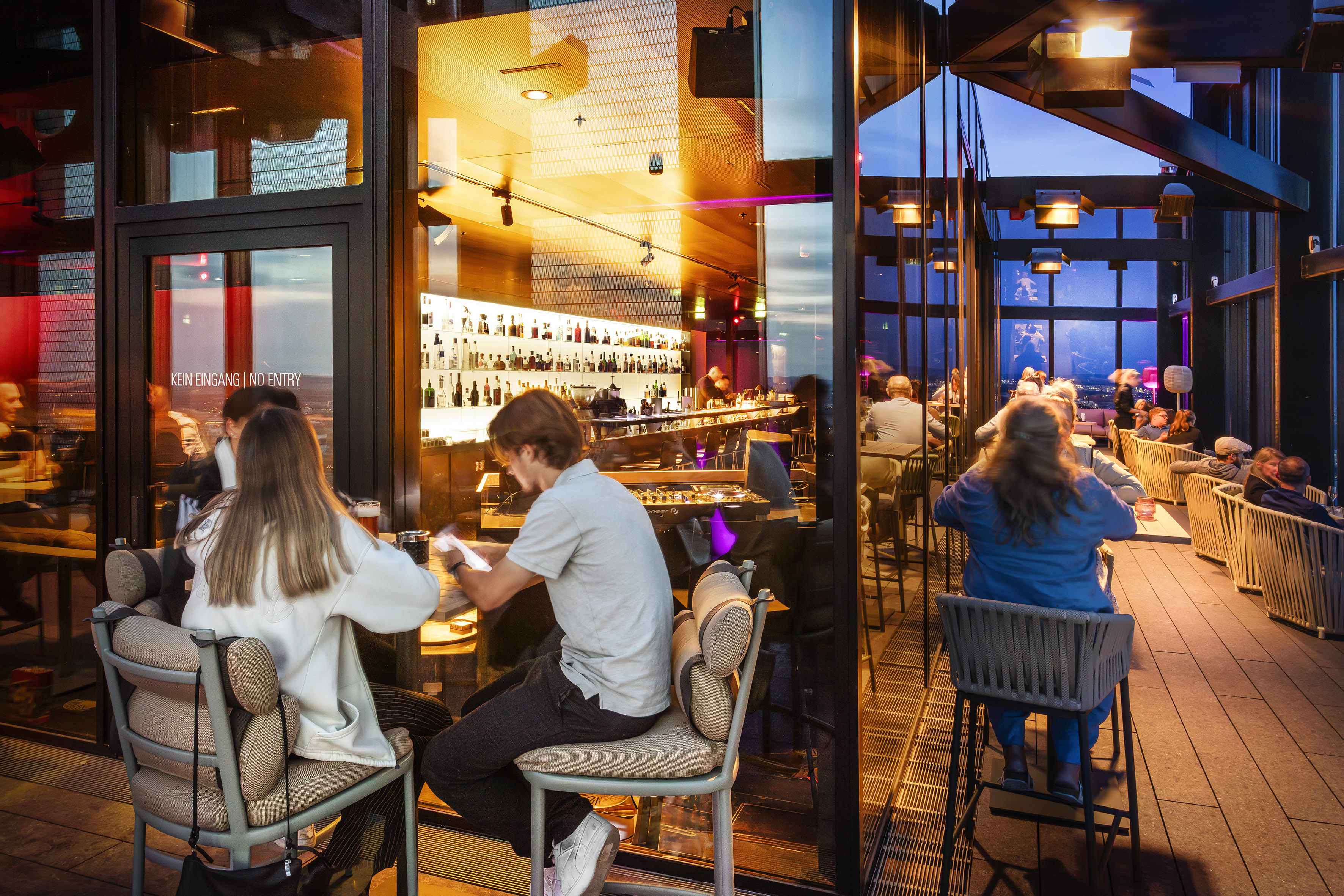 Modern rooftop bar with patrons enjoying drinks and urban evening views.