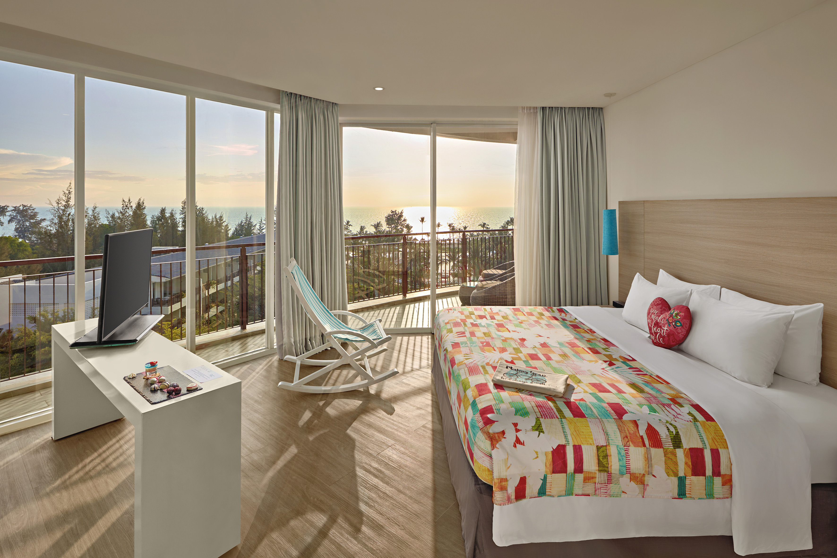 Hotel room with ocean view balcony at sunset, colorful quilt, and rocking chair.