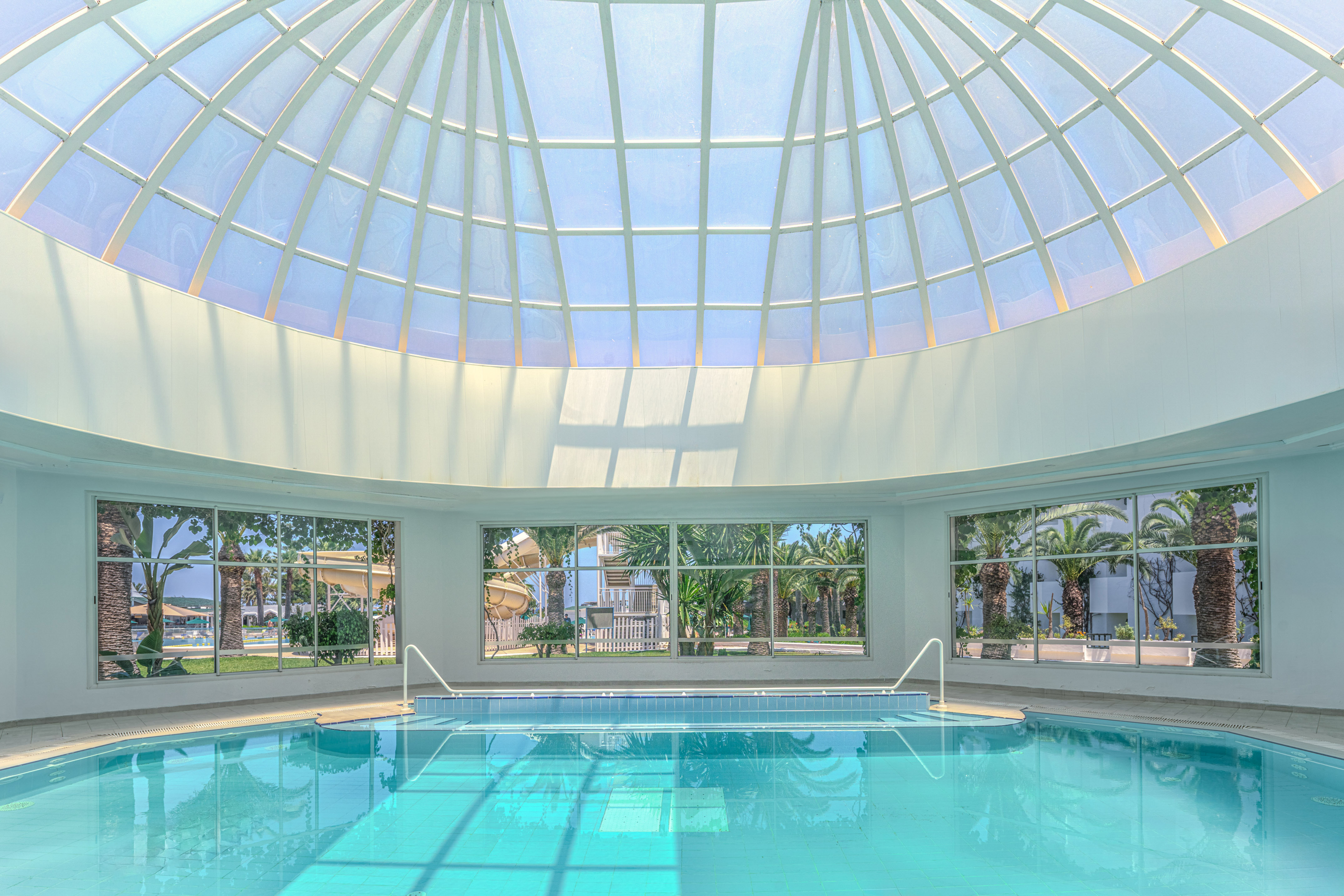 Piscina interior con cúpula de cristal, luz natural y vista a palmeras.