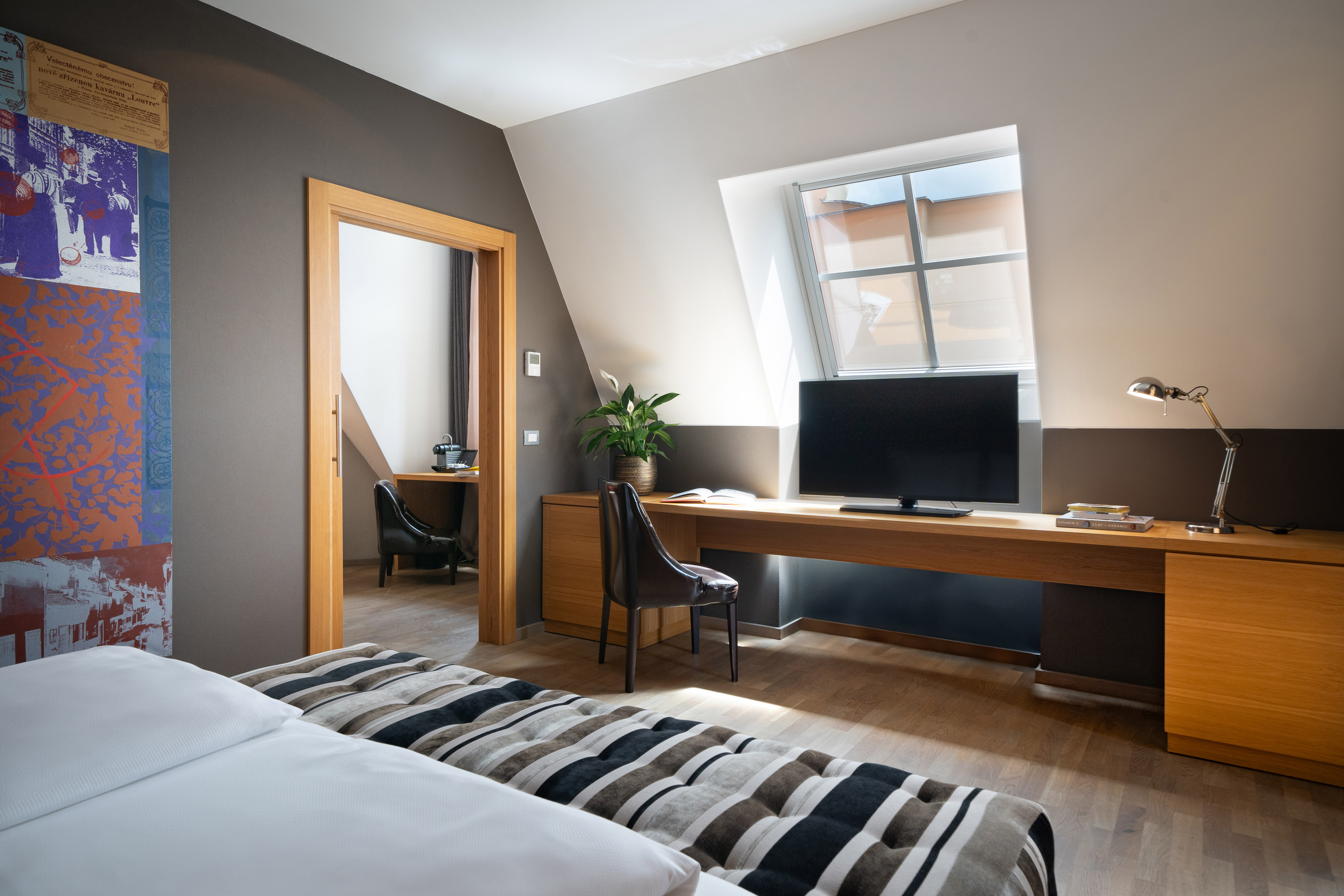 Modern hotel room with wooden desk, TV, bed, and bright attic window.