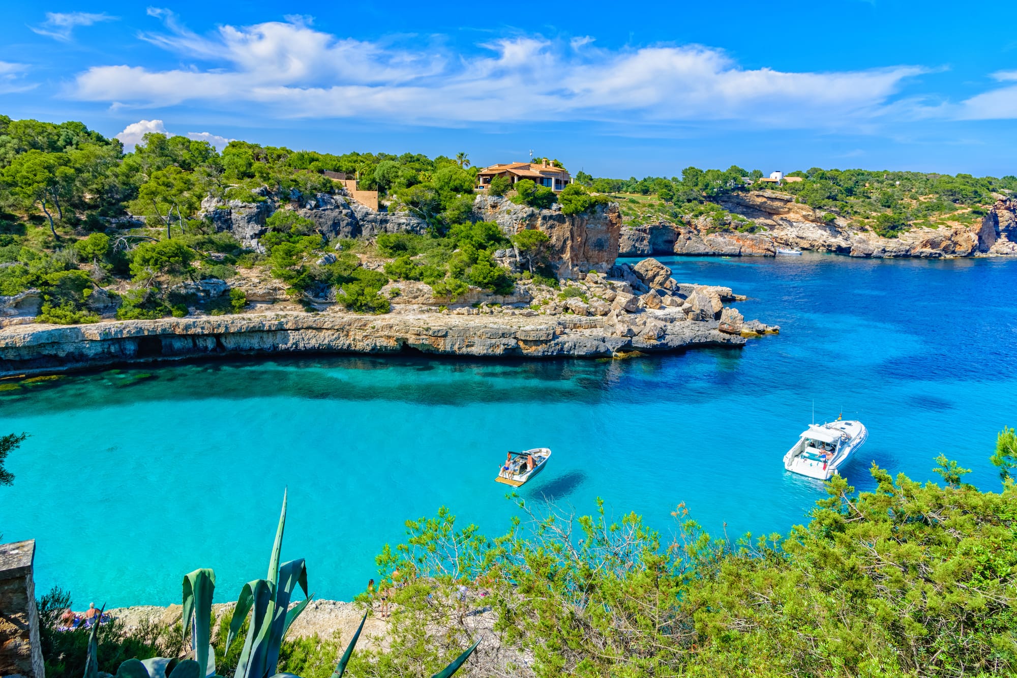 Cala Mallorca: mar turquesa, acantilados arbolados, villa y barcos.
