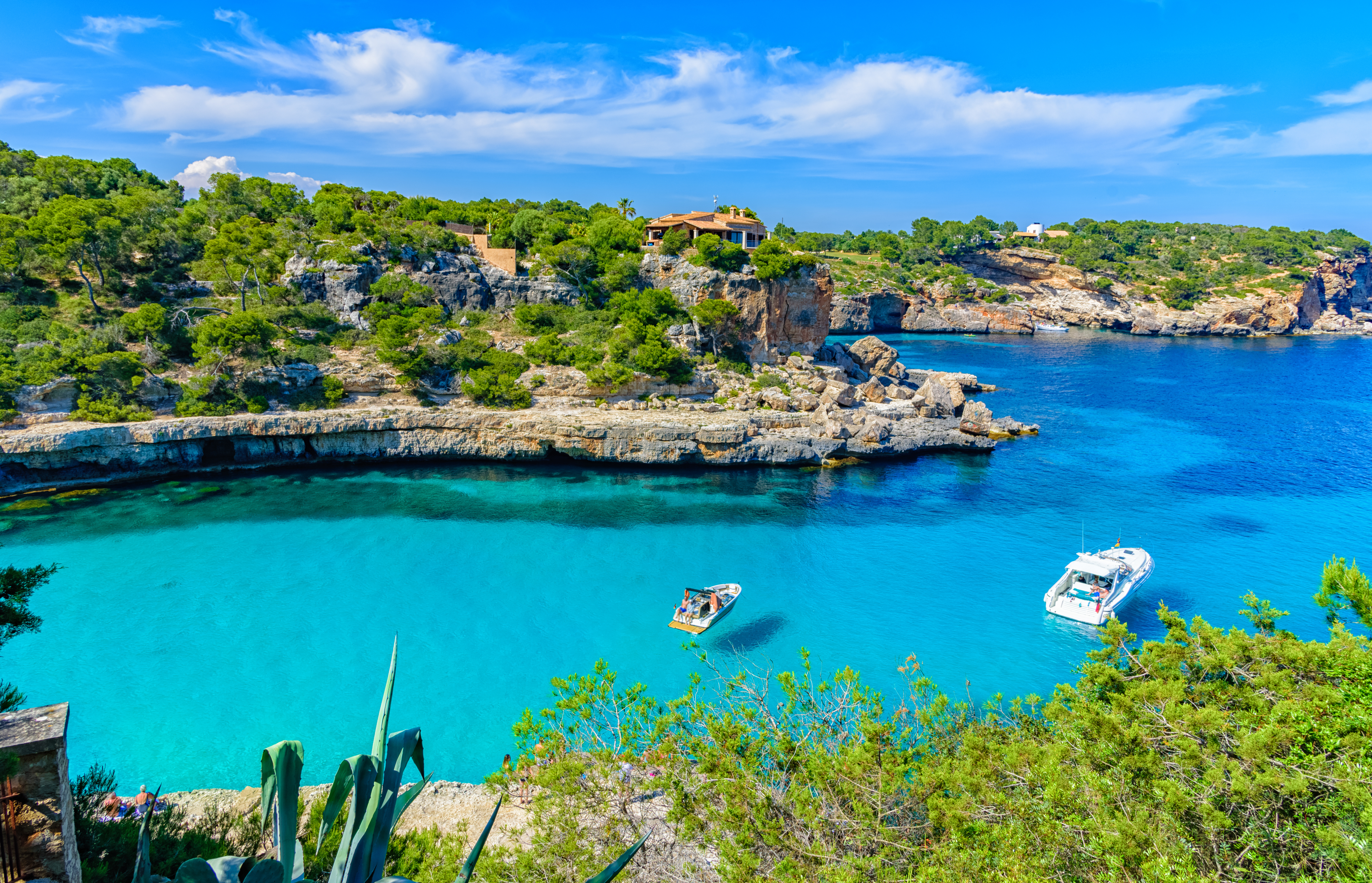 Cala Mallorca: mar turquesa, acantilados arbolados, villa y barcos.