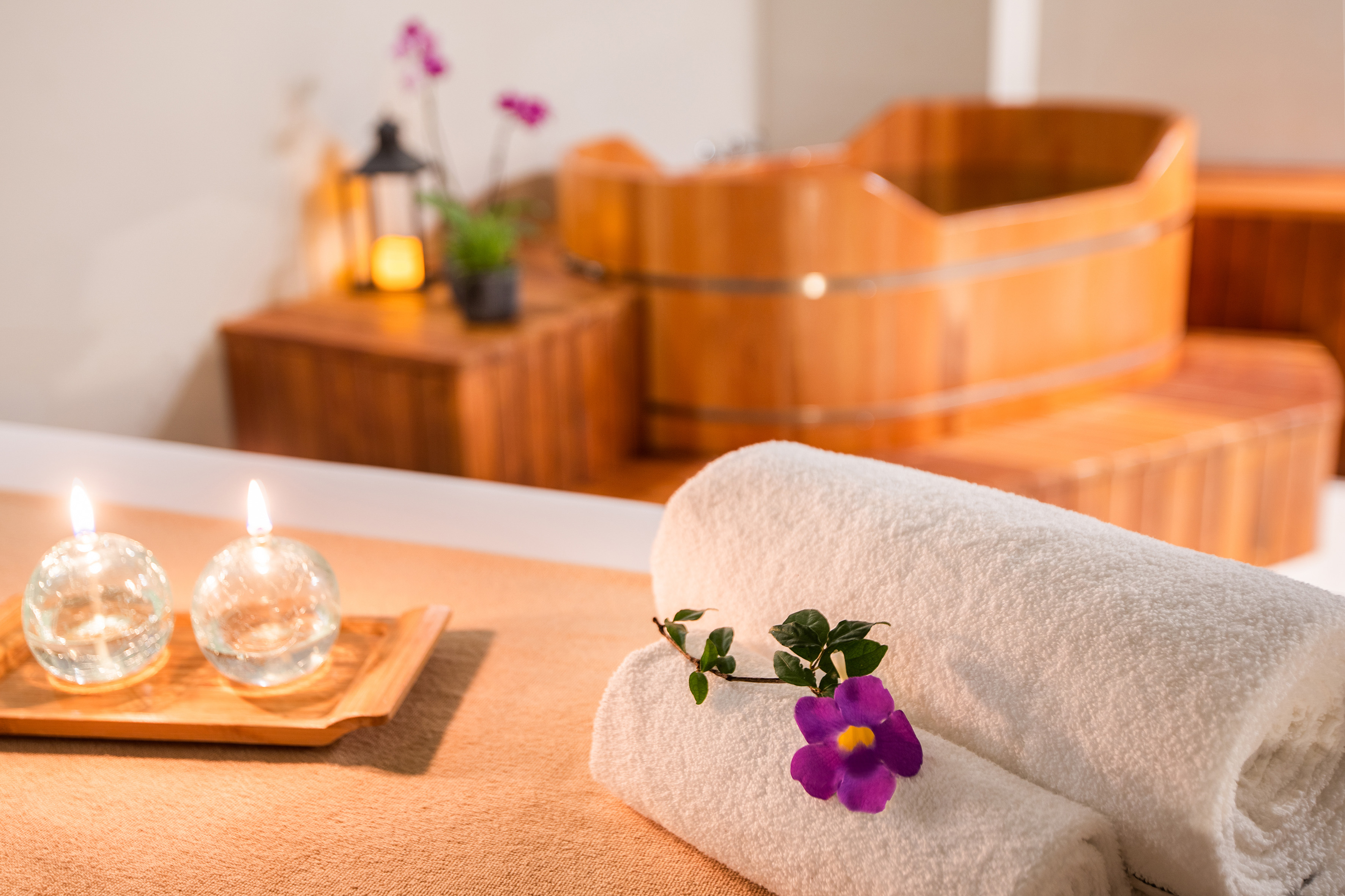 Relaxing spa bath with rolled towels, purple flower, lit candles, wooden tub.