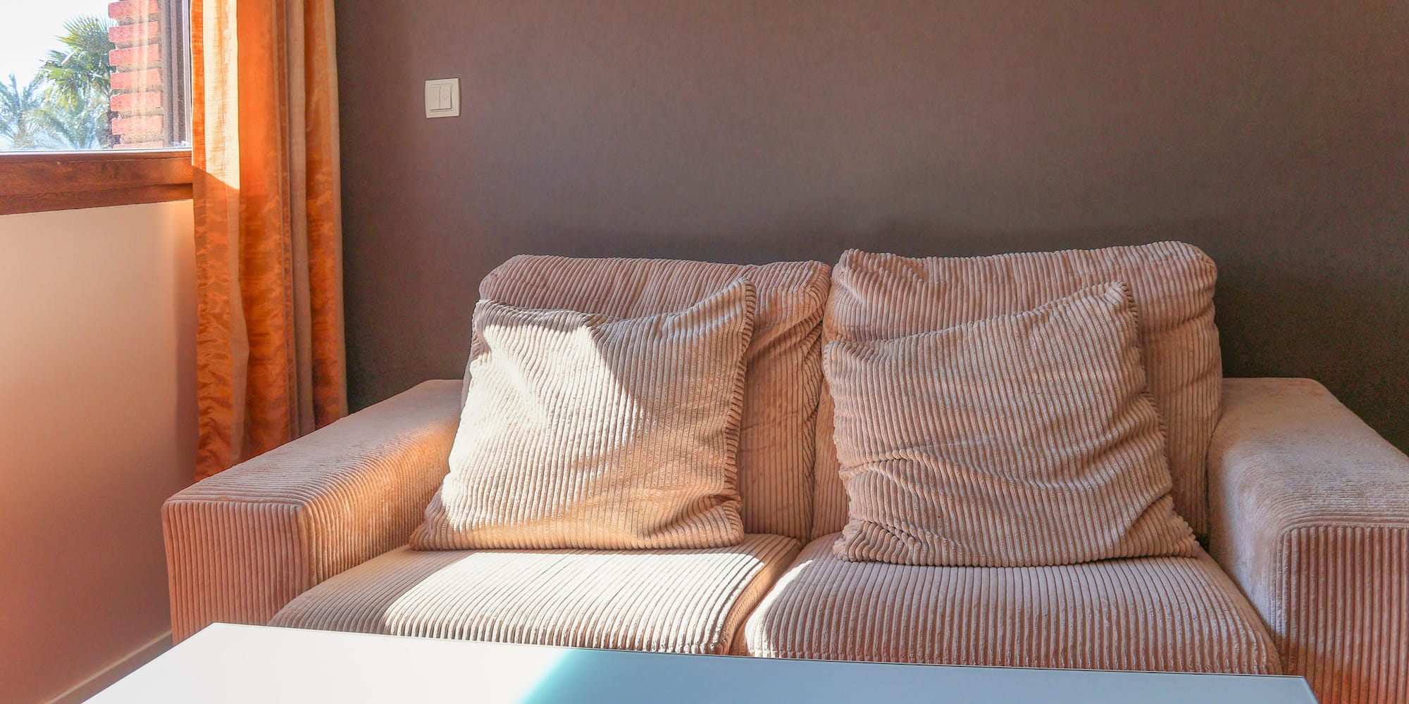 Peach corduroy sofa with pillows by a sunlit window and orange curtains.
