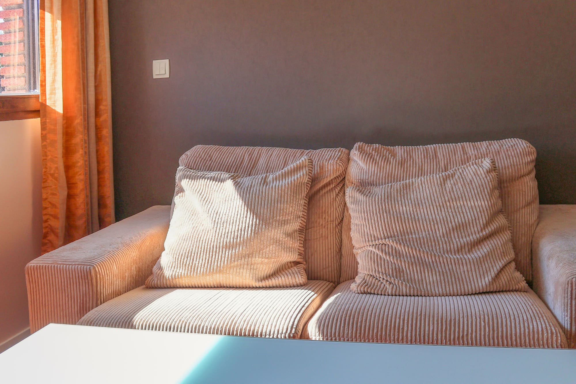Peach corduroy sofa with pillows by a sunlit window and orange curtains.