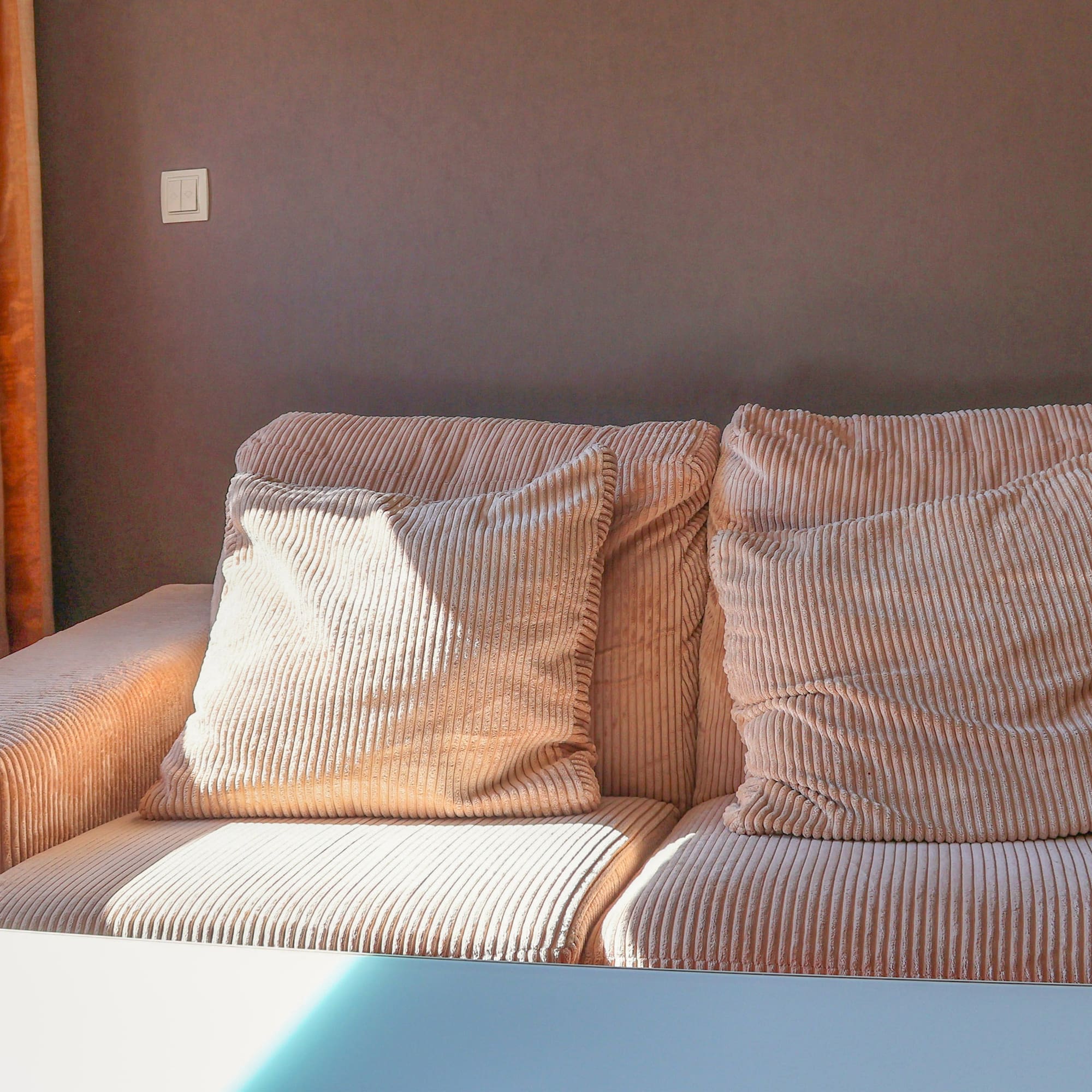 Peach corduroy sofa with pillows by a sunlit window and orange curtains.