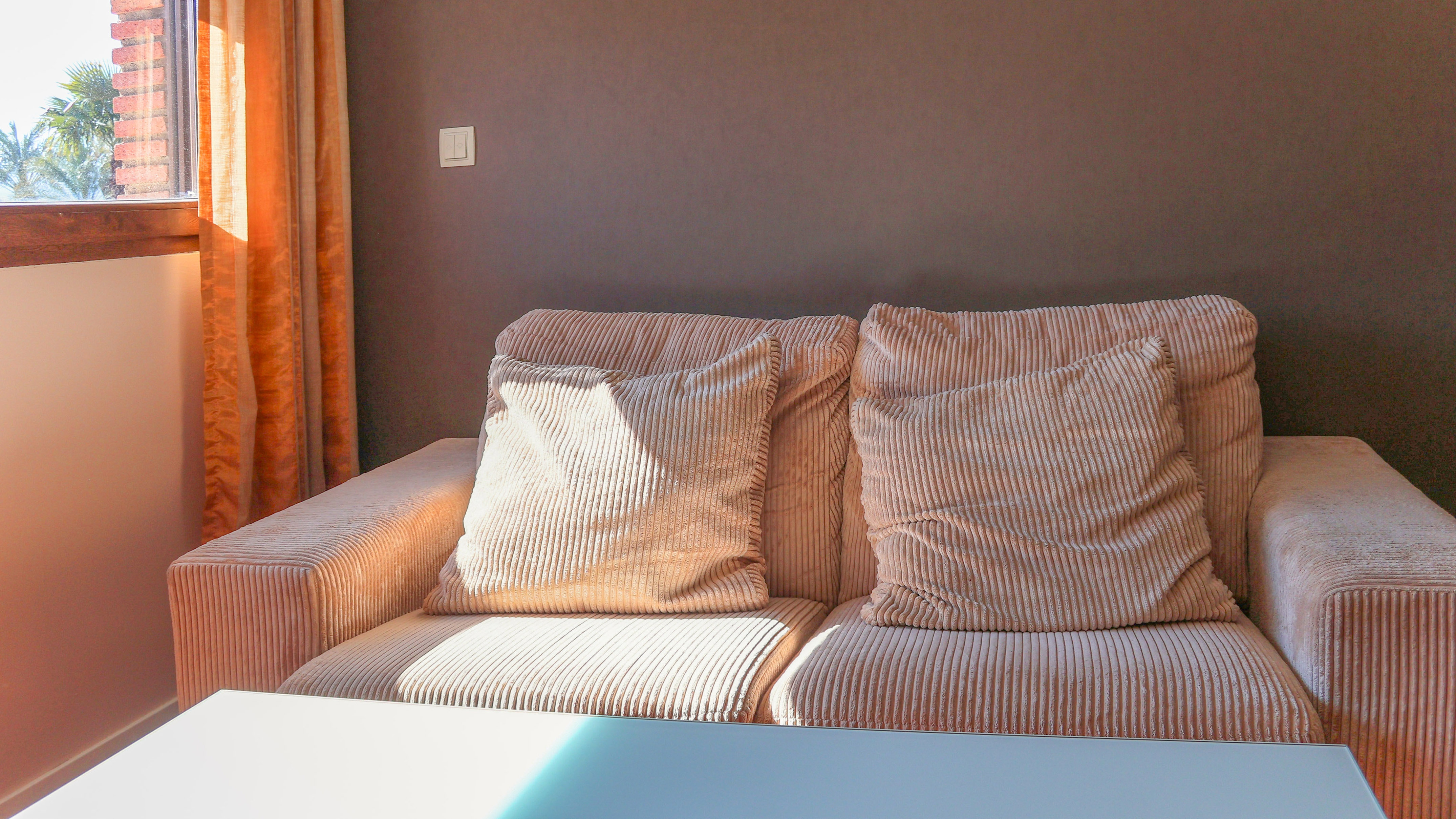 Peach corduroy sofa with pillows by a sunlit window and orange curtains.