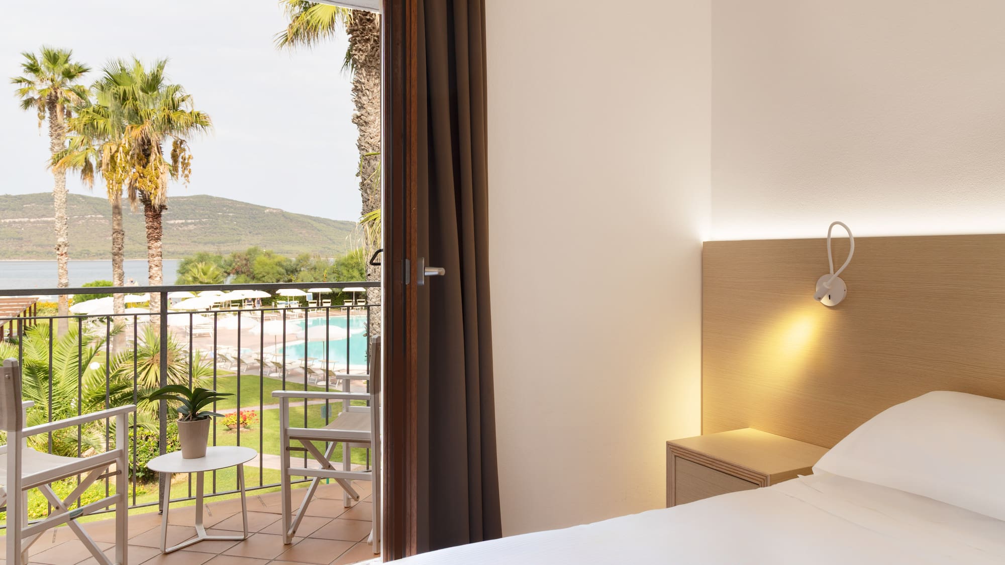 Hotel room with balcony overlooking pool, sea, and palm trees.