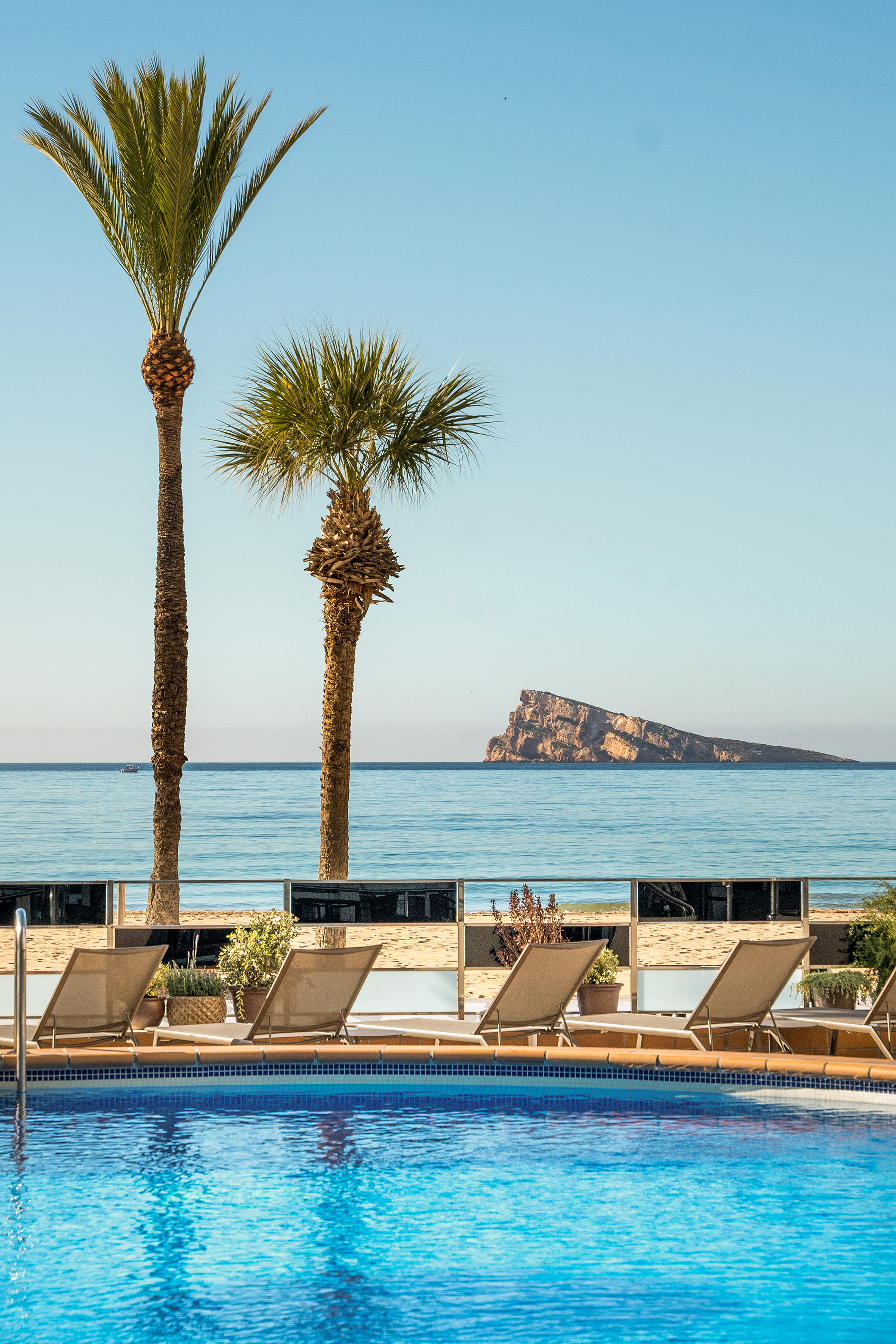 Clear blue pool with sun loungers, palm trees, ocean, and distant rocky island.