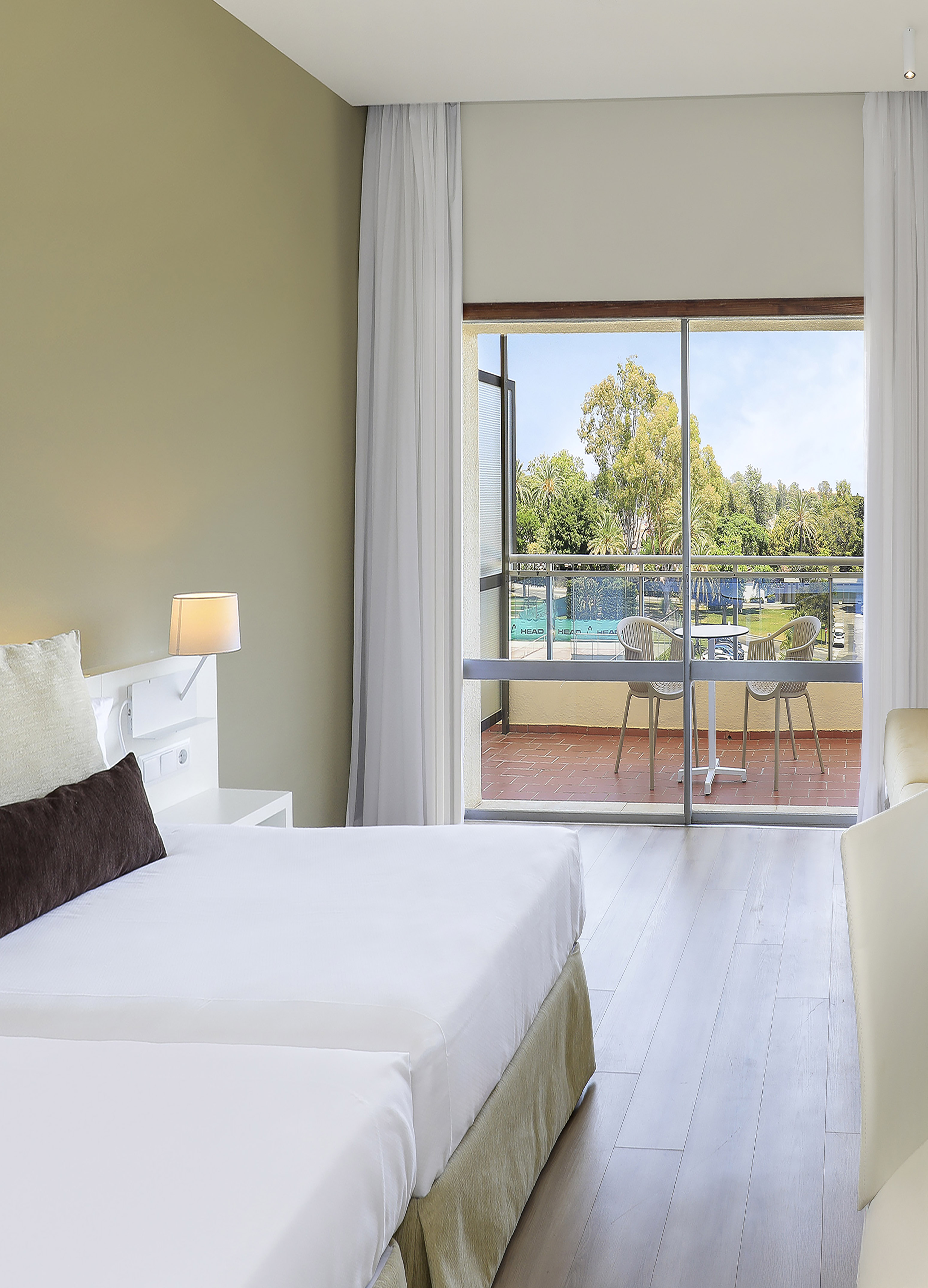 Bright hotel room with twin beds and balcony view of tennis court.