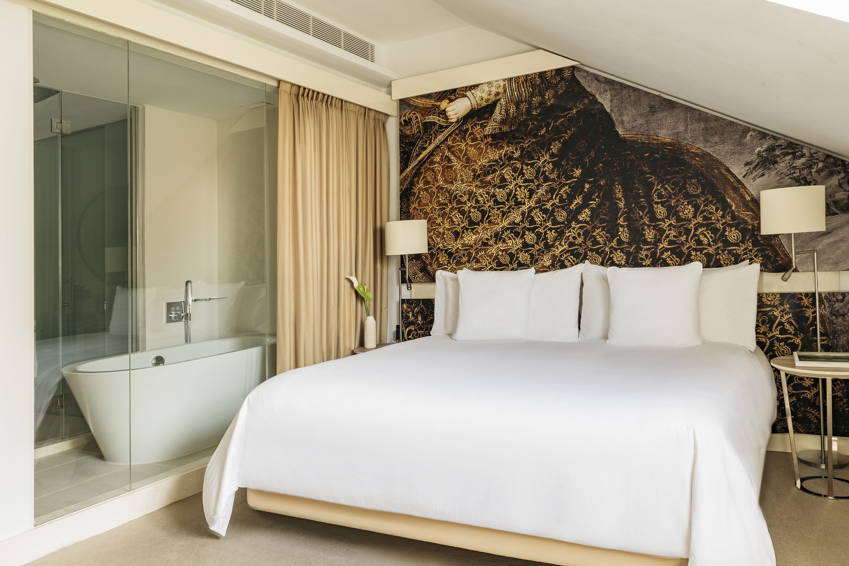 Hotel bedroom with king bed, ornate gold wallpaper, and glass-enclosed bathtub.