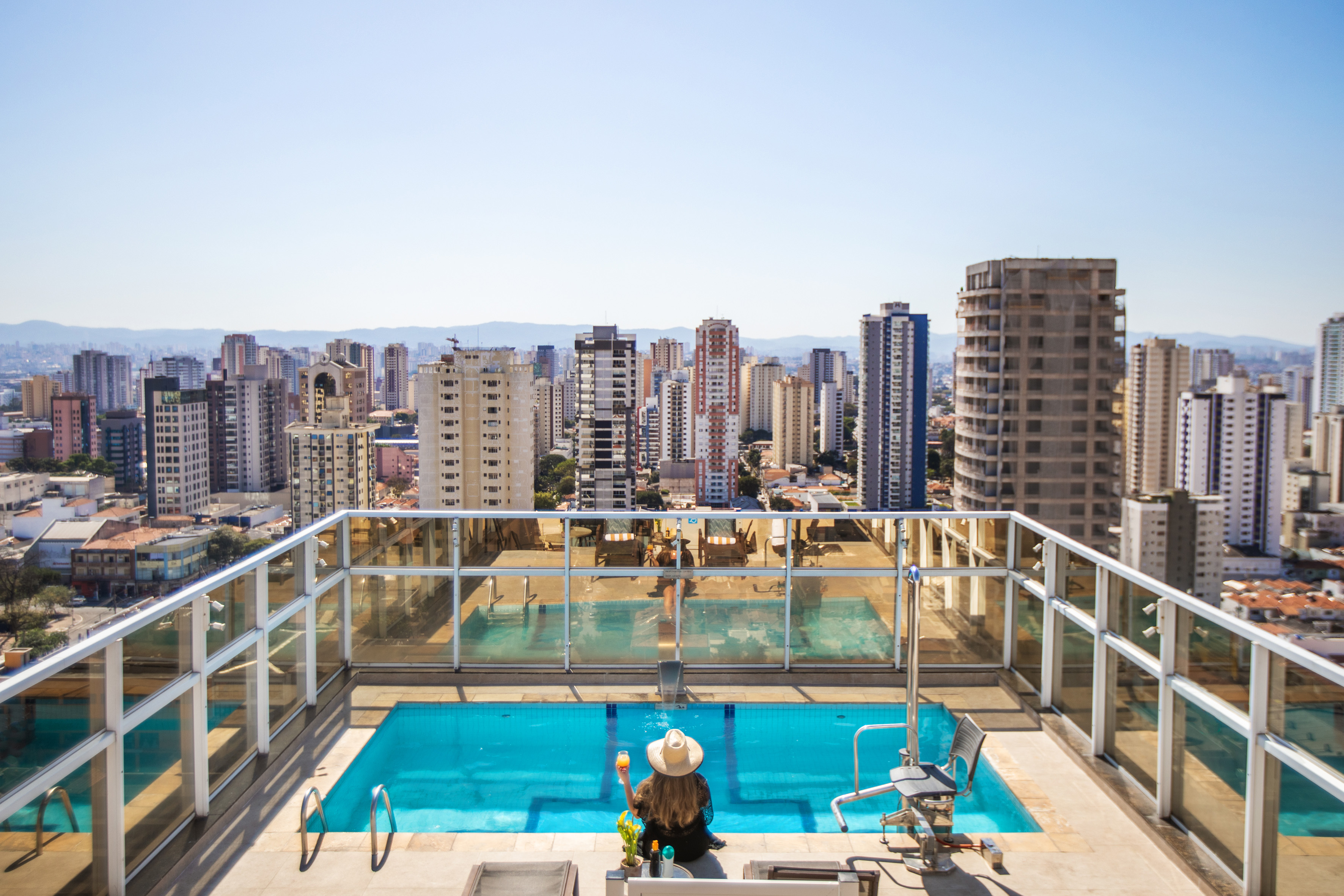 Woman enjoying a cocktail by rooftop pool with expansive city skyline view.
