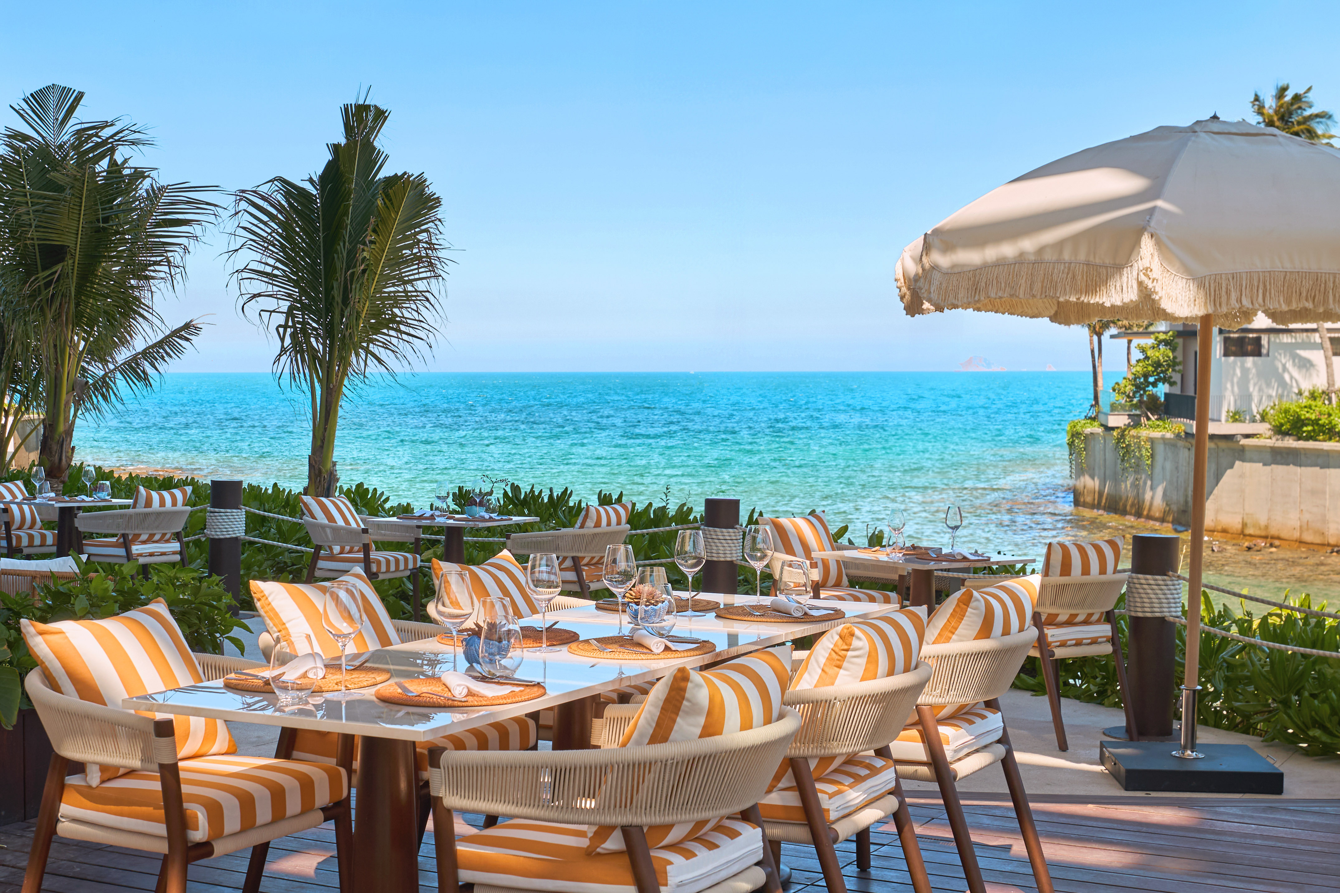 Beachfront restaurant with orange chairs, palm trees, and clear blue sea.
