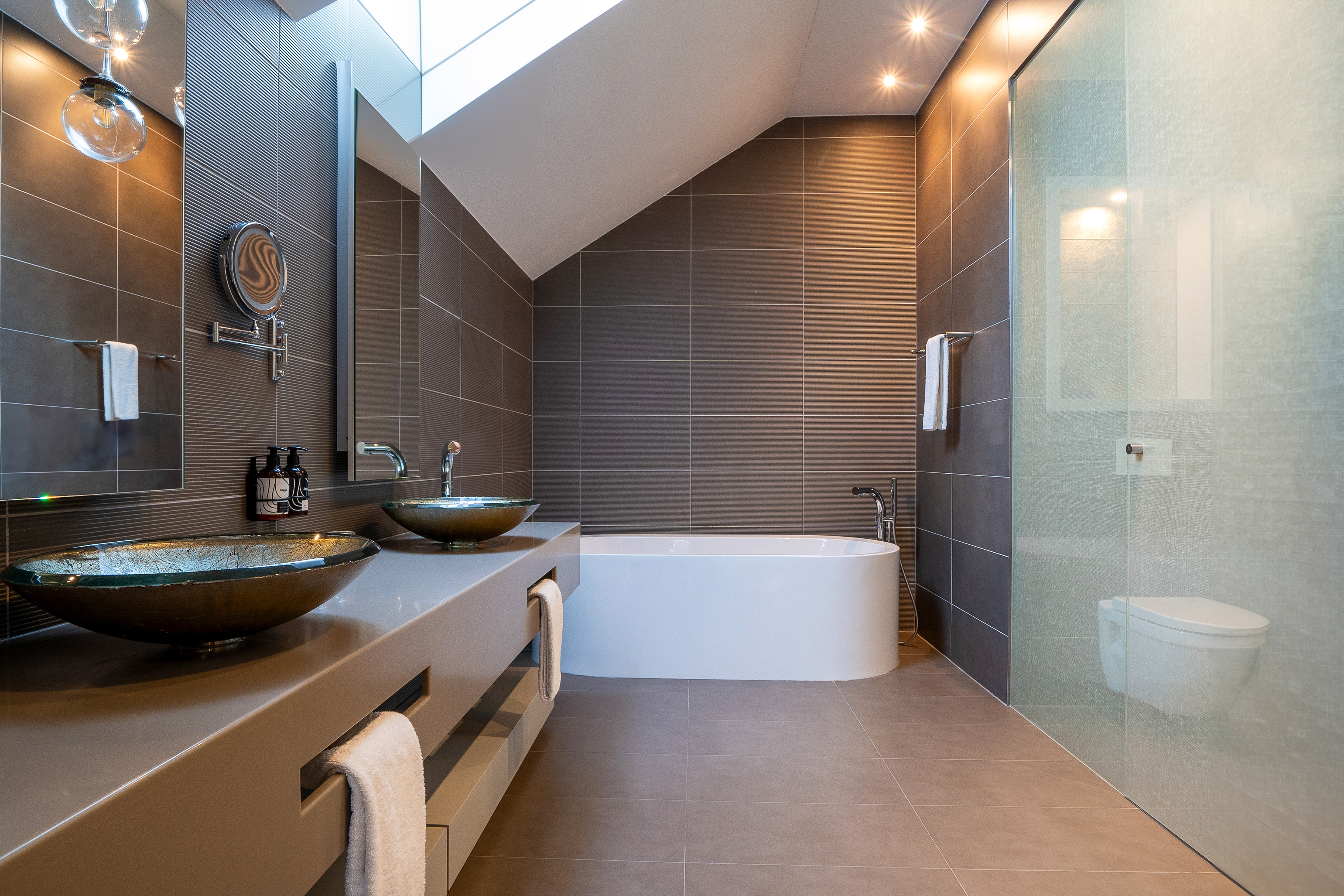 Modern bathroom with freestanding tub, vessel sinks, dark tiles, and skylight.