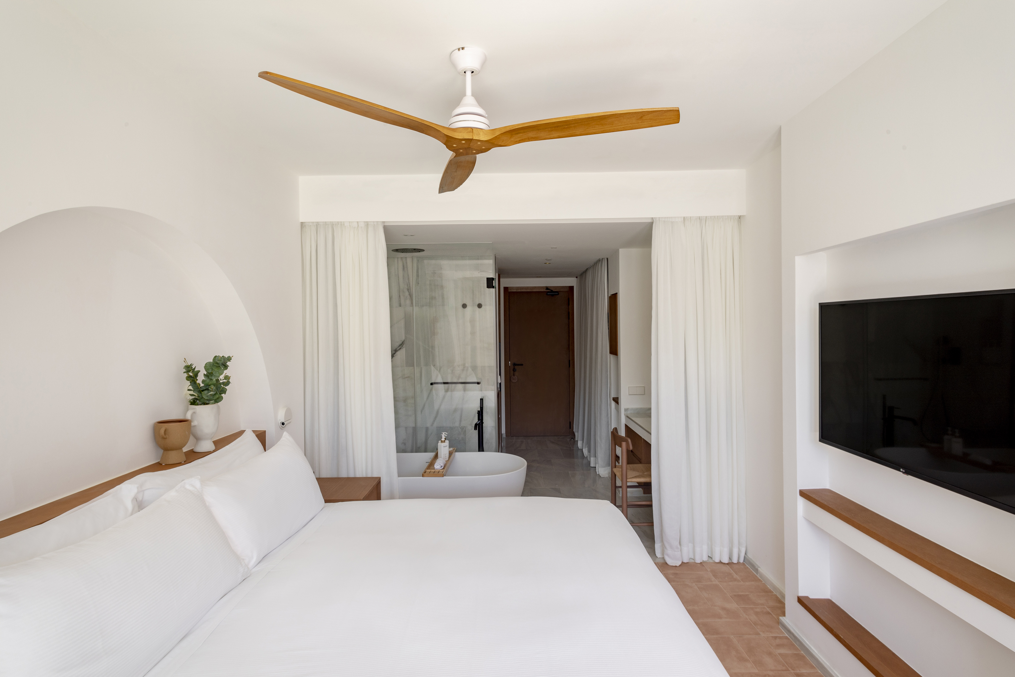 Modern bedroom with white bed, wooden fan, and open bathroom with freestanding tub.