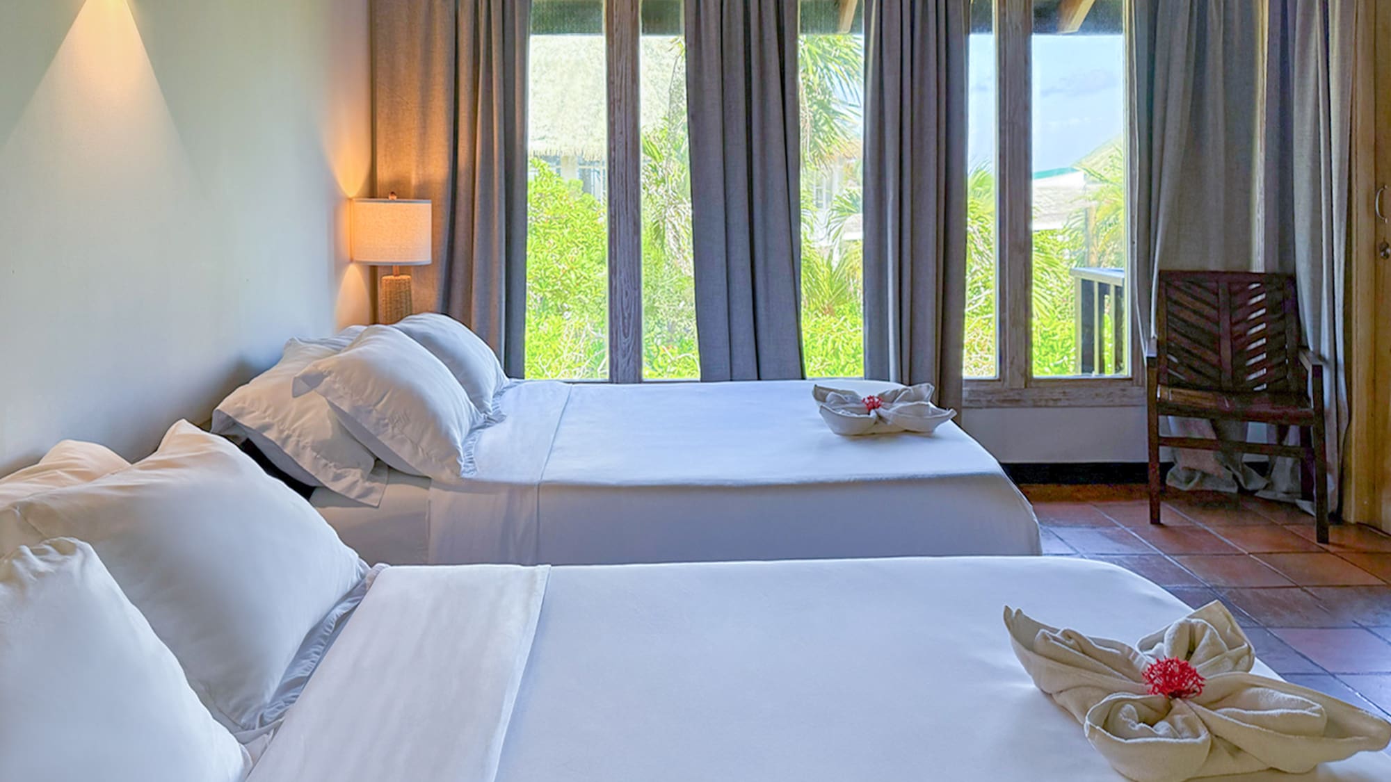 Two beds with white bedding in a hotel room with tropical view windows.