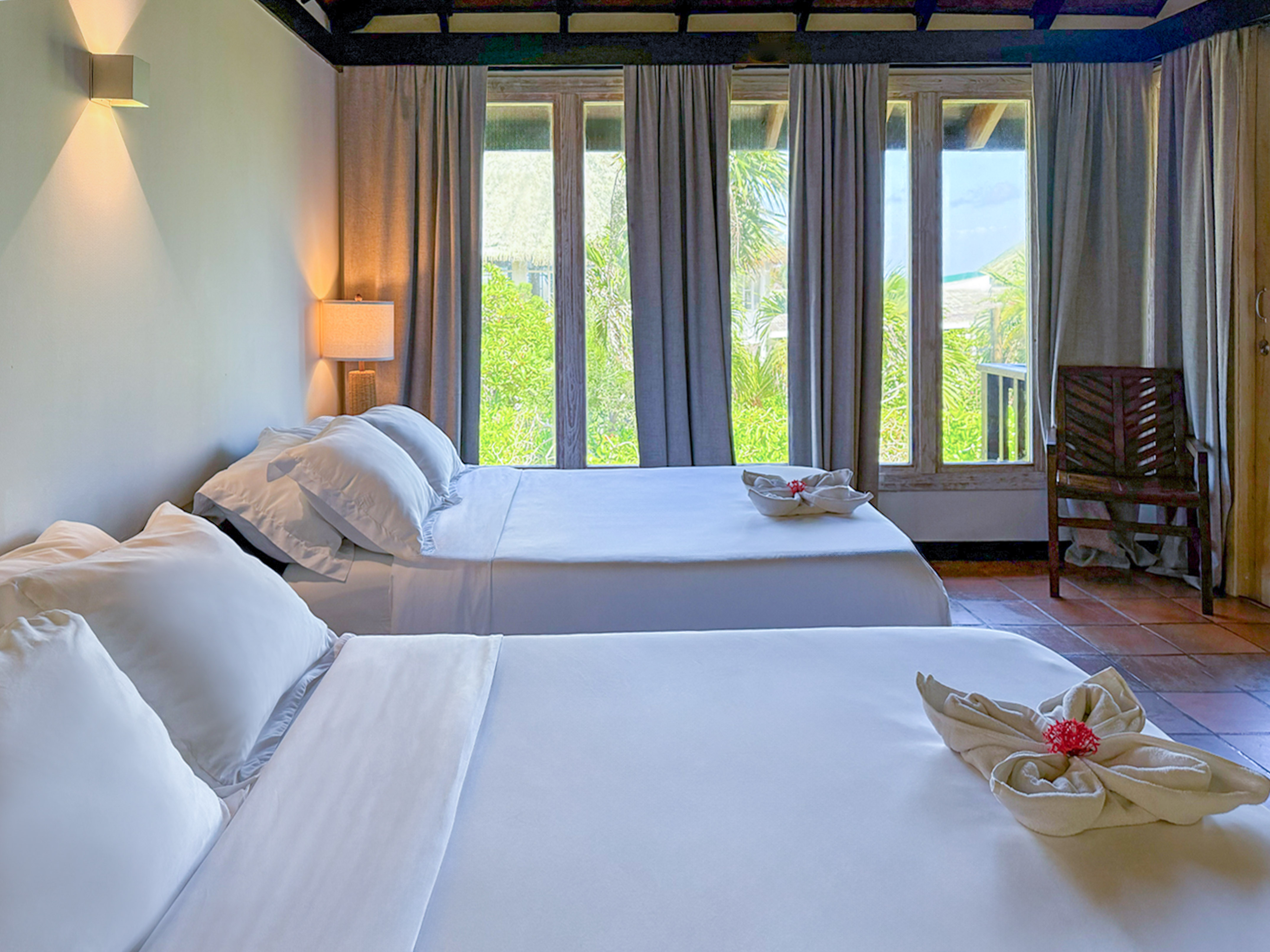 Two beds with white bedding in a hotel room with tropical view windows.