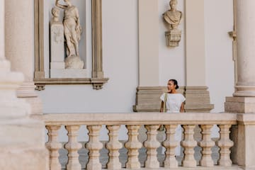 Woman in white dress on classical stone balcony with statues.