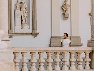 Woman in white dress on classical stone balcony with statues.