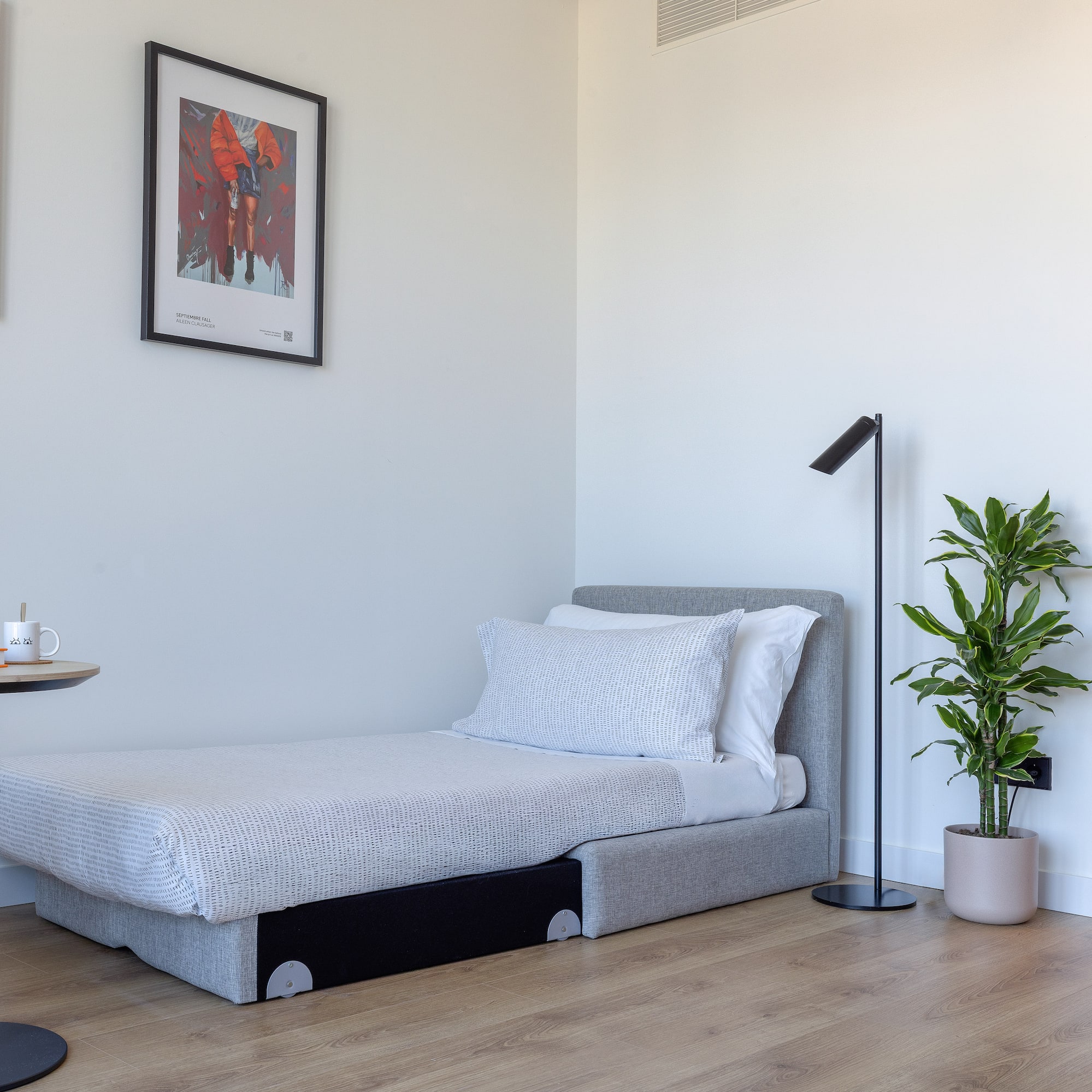 Estancia moderna con cama individual, mesa auxiliar, planta y arte en pared.