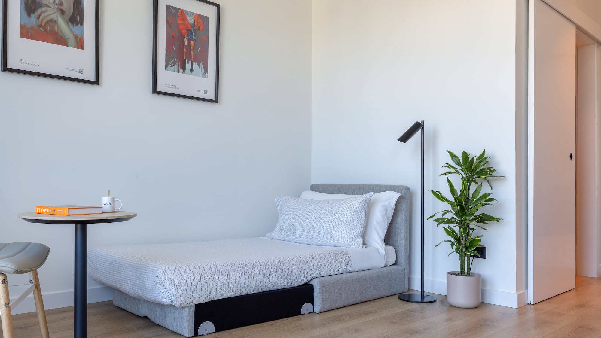 Estancia moderna con cama individual, mesa auxiliar, planta y arte en pared.