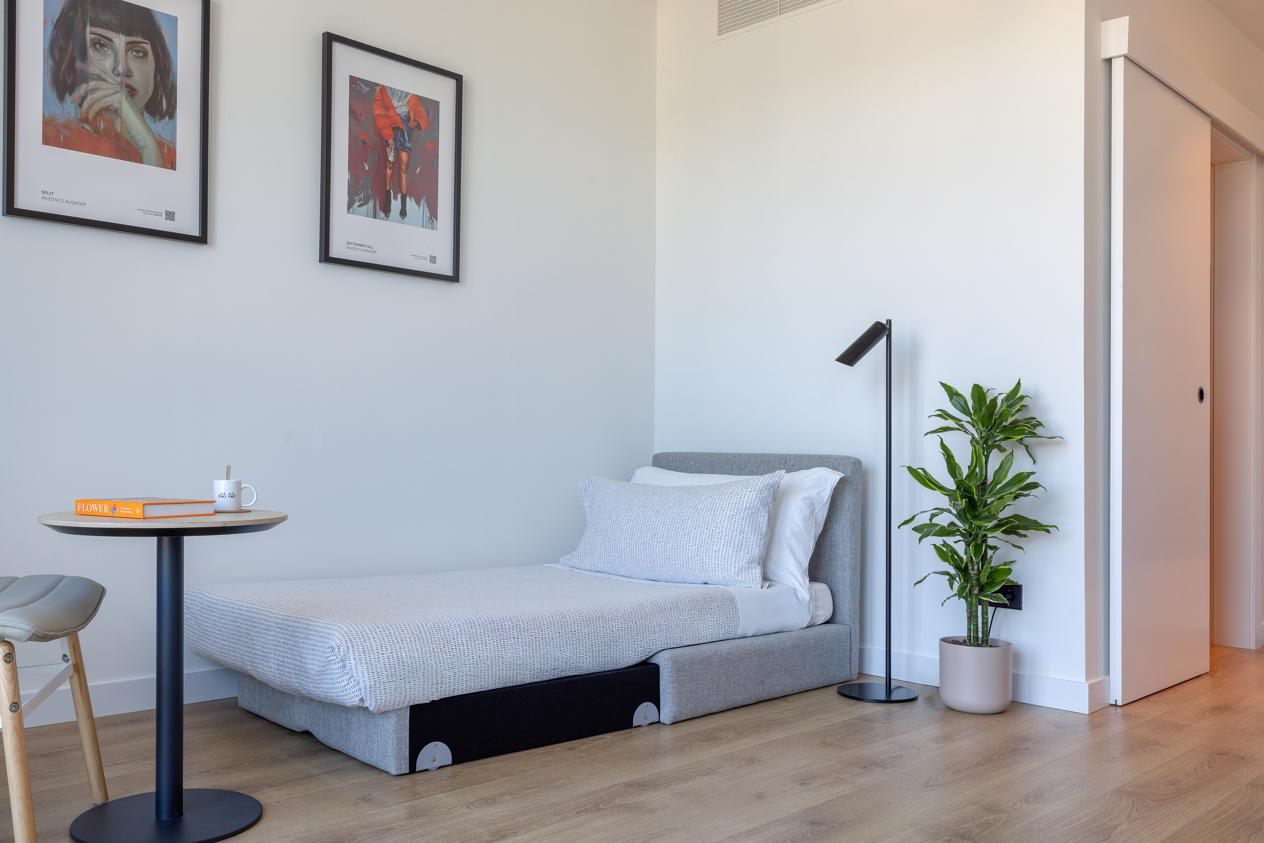 Estancia moderna con cama individual, mesa auxiliar, planta y arte en pared.