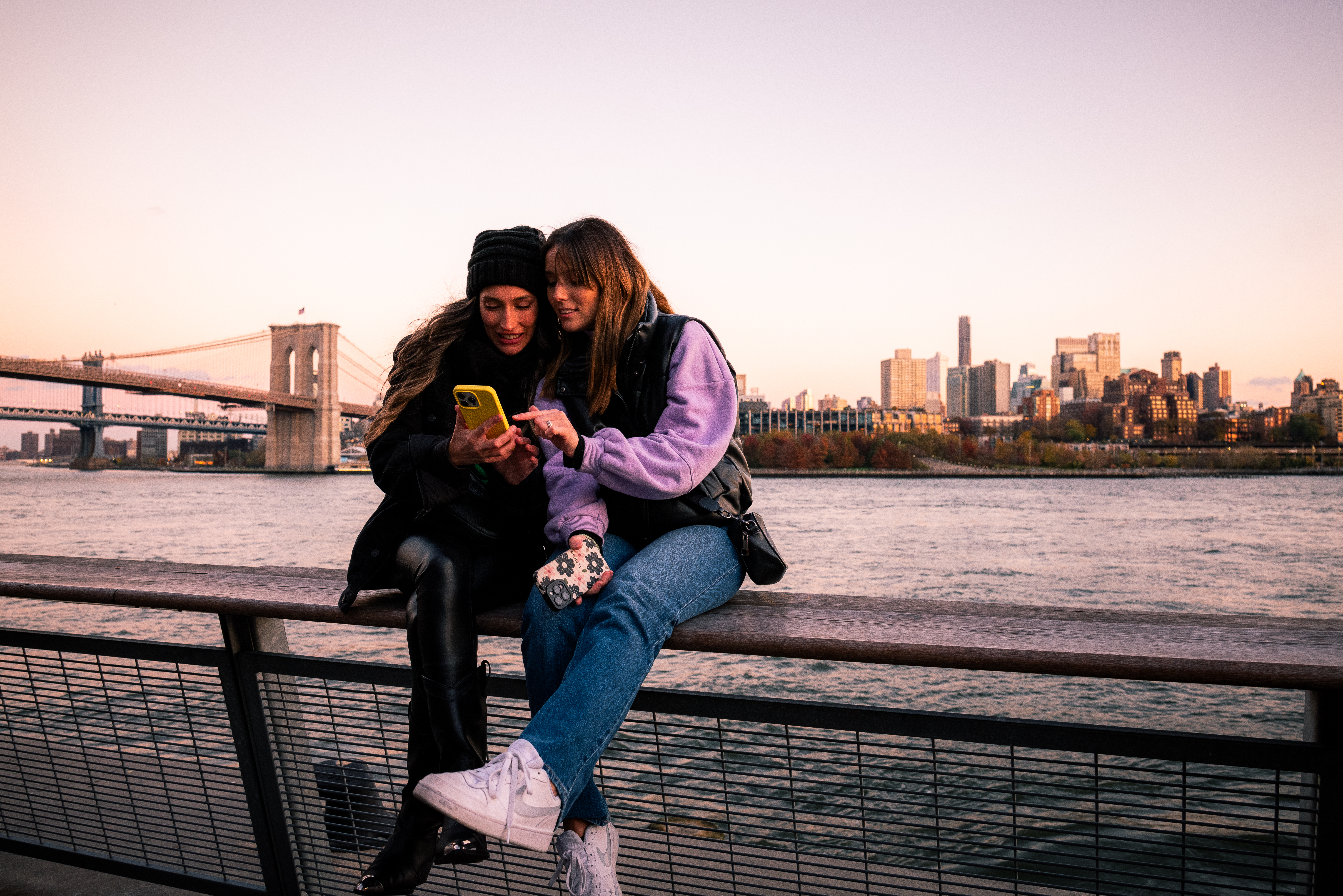 two women sitting on a railing looking at a phone
