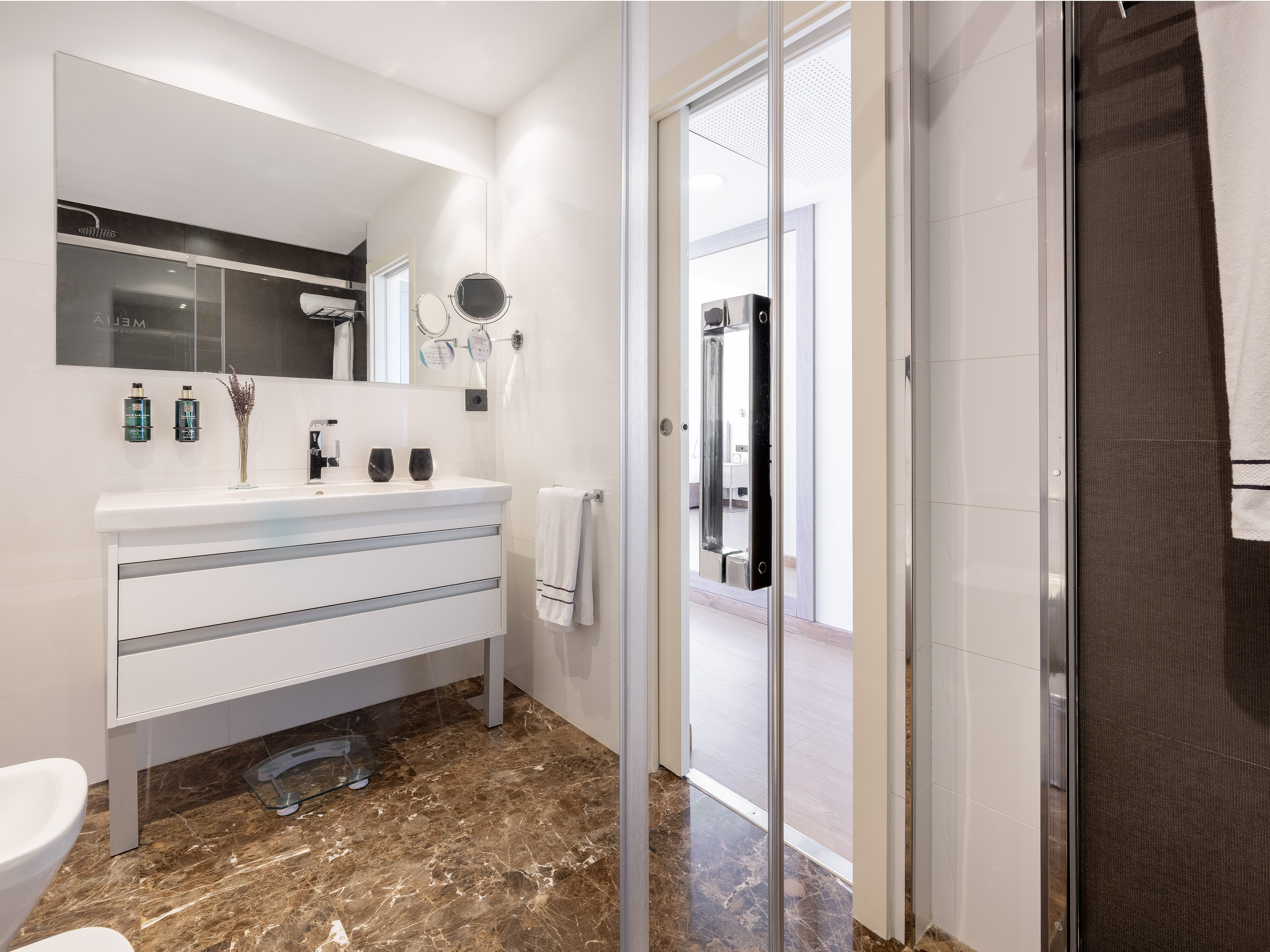 Modern bathroom with white vanity, mirror, glass shower, brown marble floor.