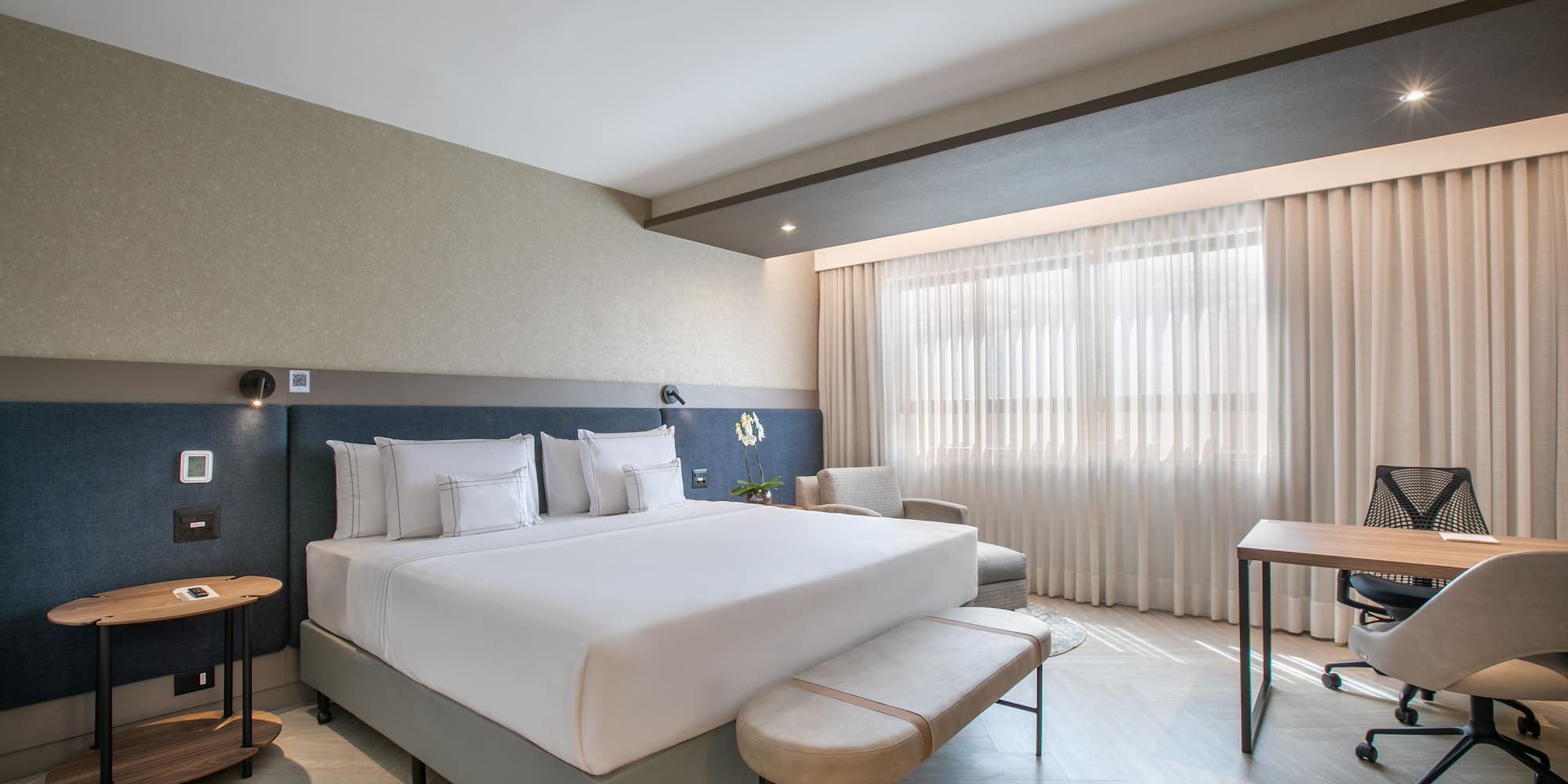 Modern hotel bedroom with king bed, nightstand, desk, and bright window.