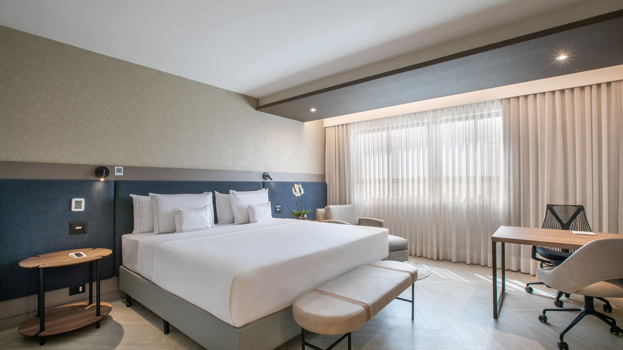 Modern hotel bedroom with king bed, nightstand, desk, and bright window.