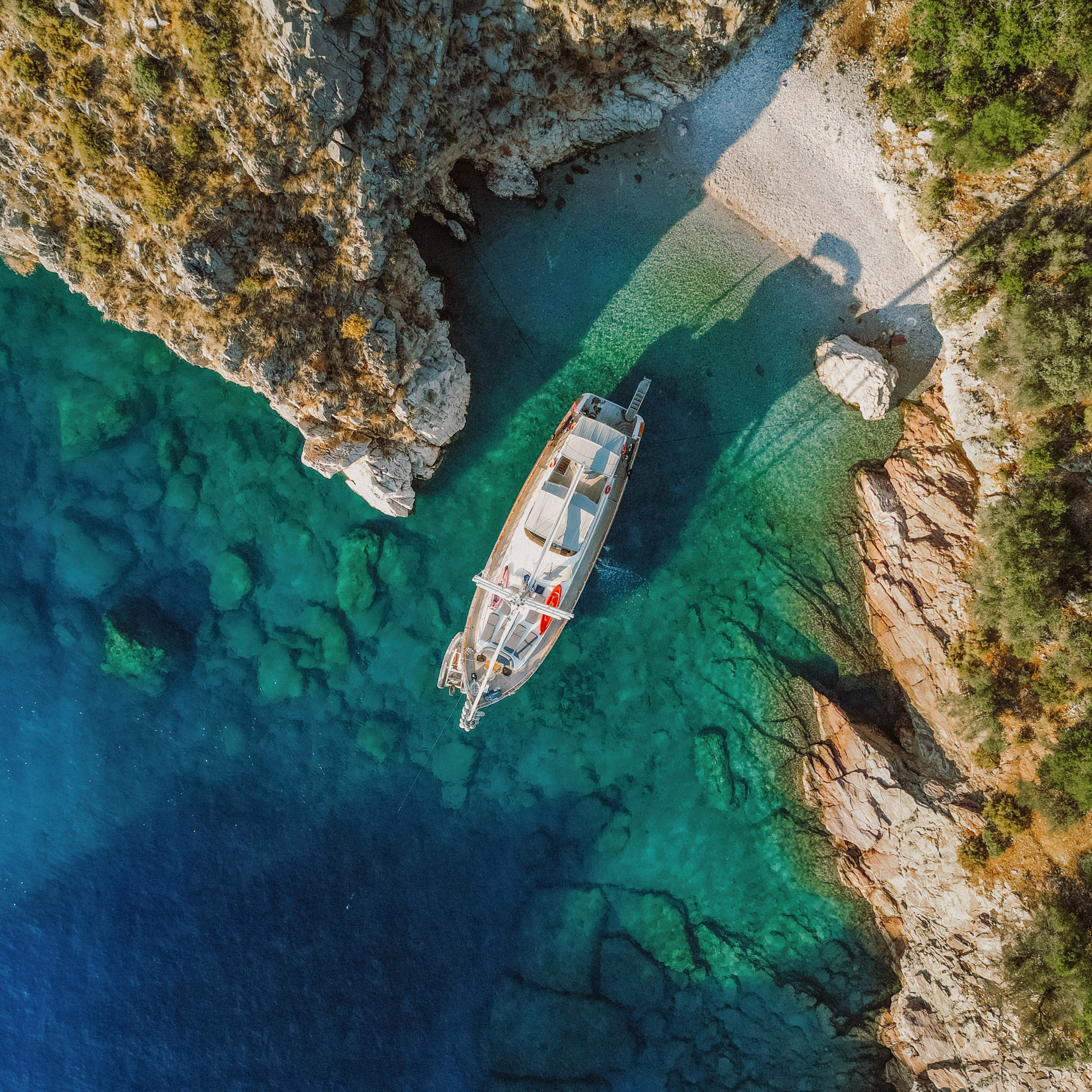Aerial view of yacht in clear turquoise bay with rocky cliffs and beach.