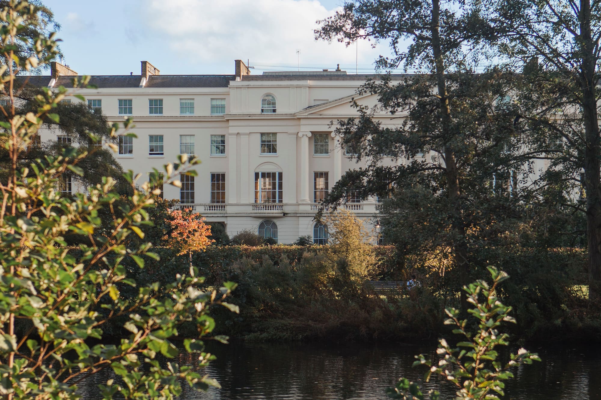 Grand neoclassical building overlooking a reflective lake, framed by autumnal trees.