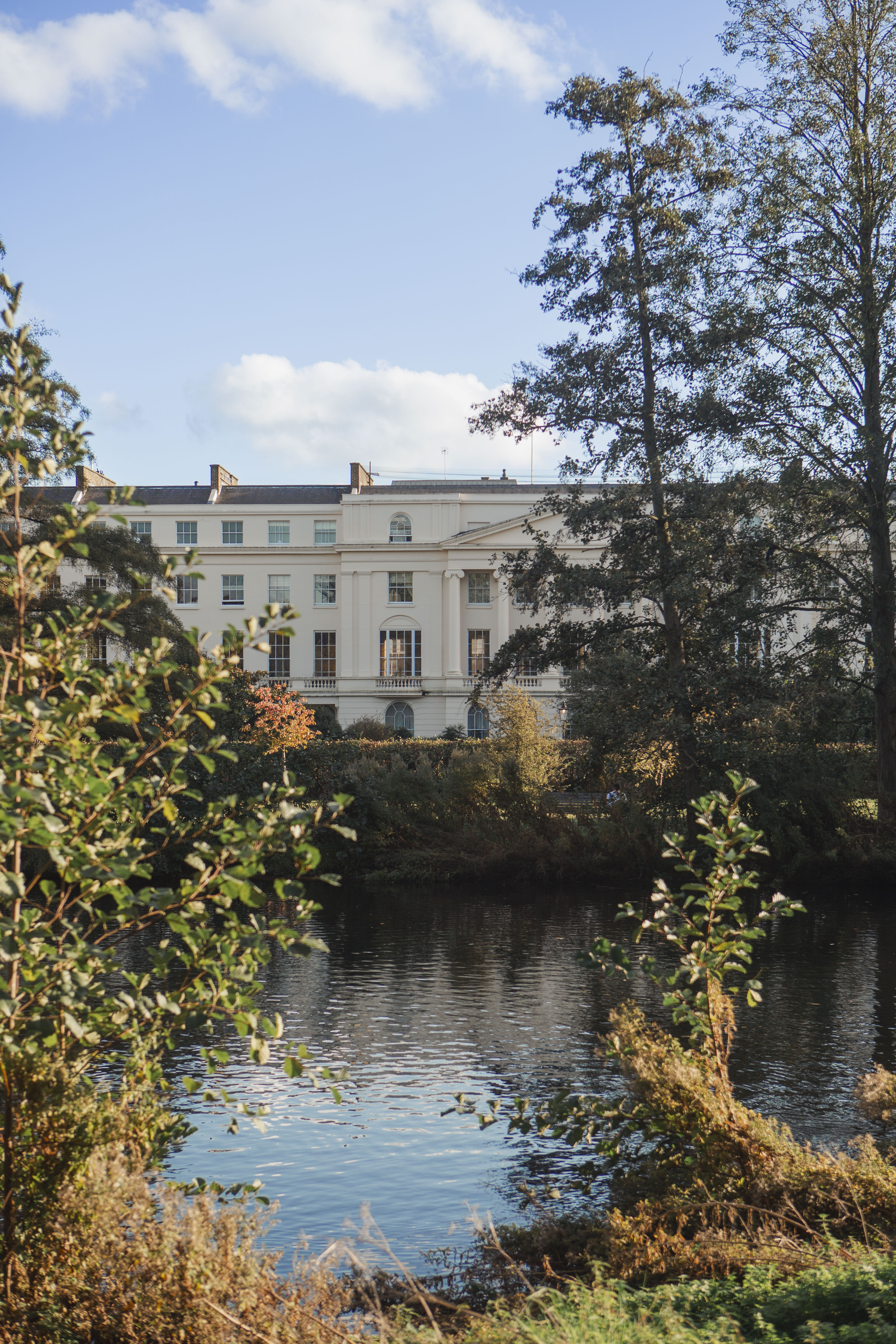 Grand neoclassical building overlooking a reflective lake, framed by autumnal trees.