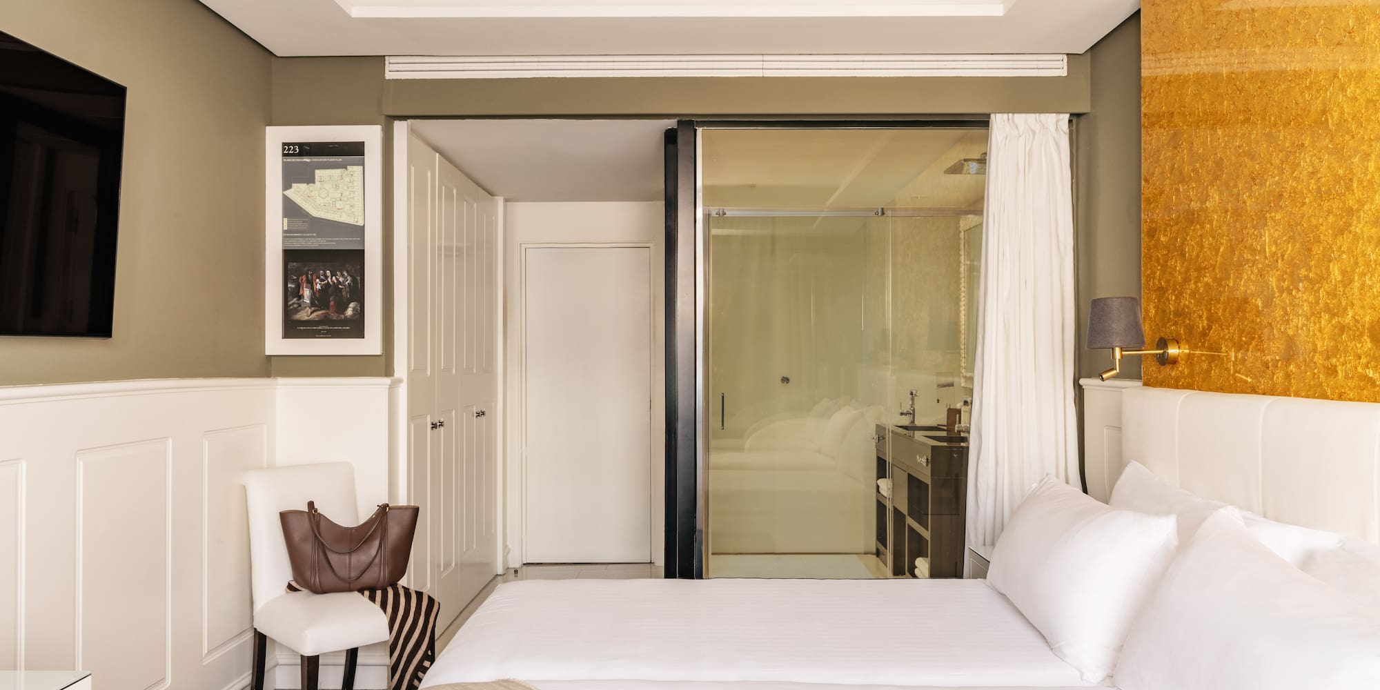 Modern hotel bedroom with twin beds, gold accent wall, and glass bathroom.