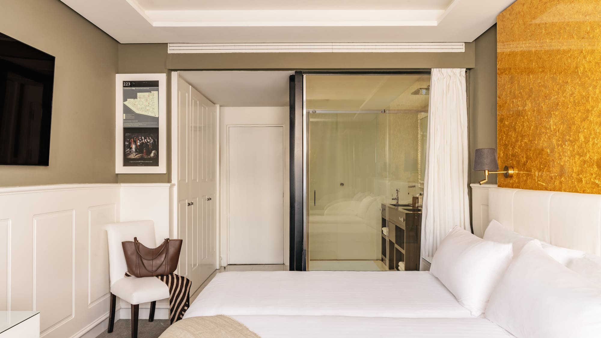 Modern hotel bedroom with twin beds, gold accent wall, and glass bathroom.