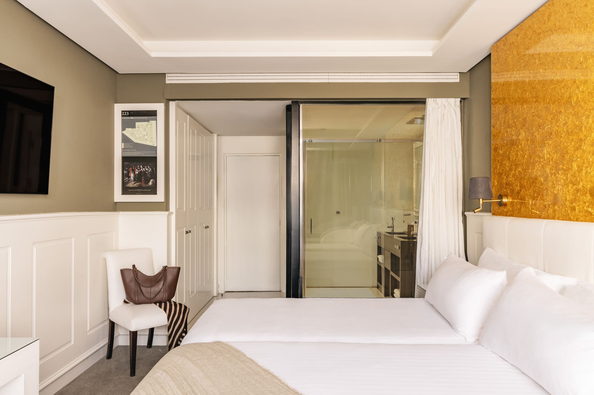 Modern hotel bedroom with twin beds, gold accent wall, and glass bathroom.